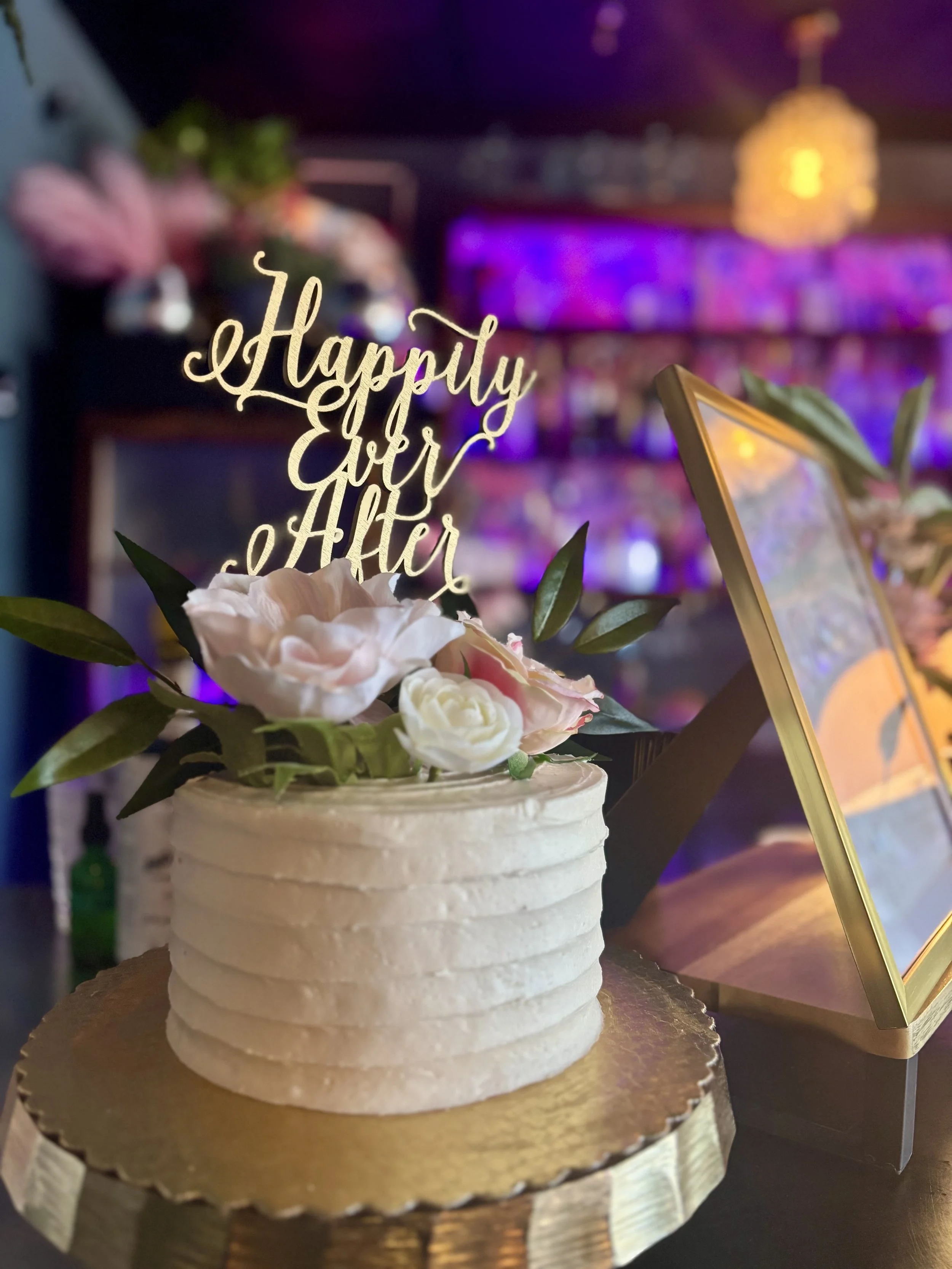 White cake with floral decorations and "Happily Ever After" topper on a gold stand, in a dimly lit setting. Baby shower, wedding shower, wedding, celebration, private dining, private events, birthdays, custom menu. restaurant rentals.