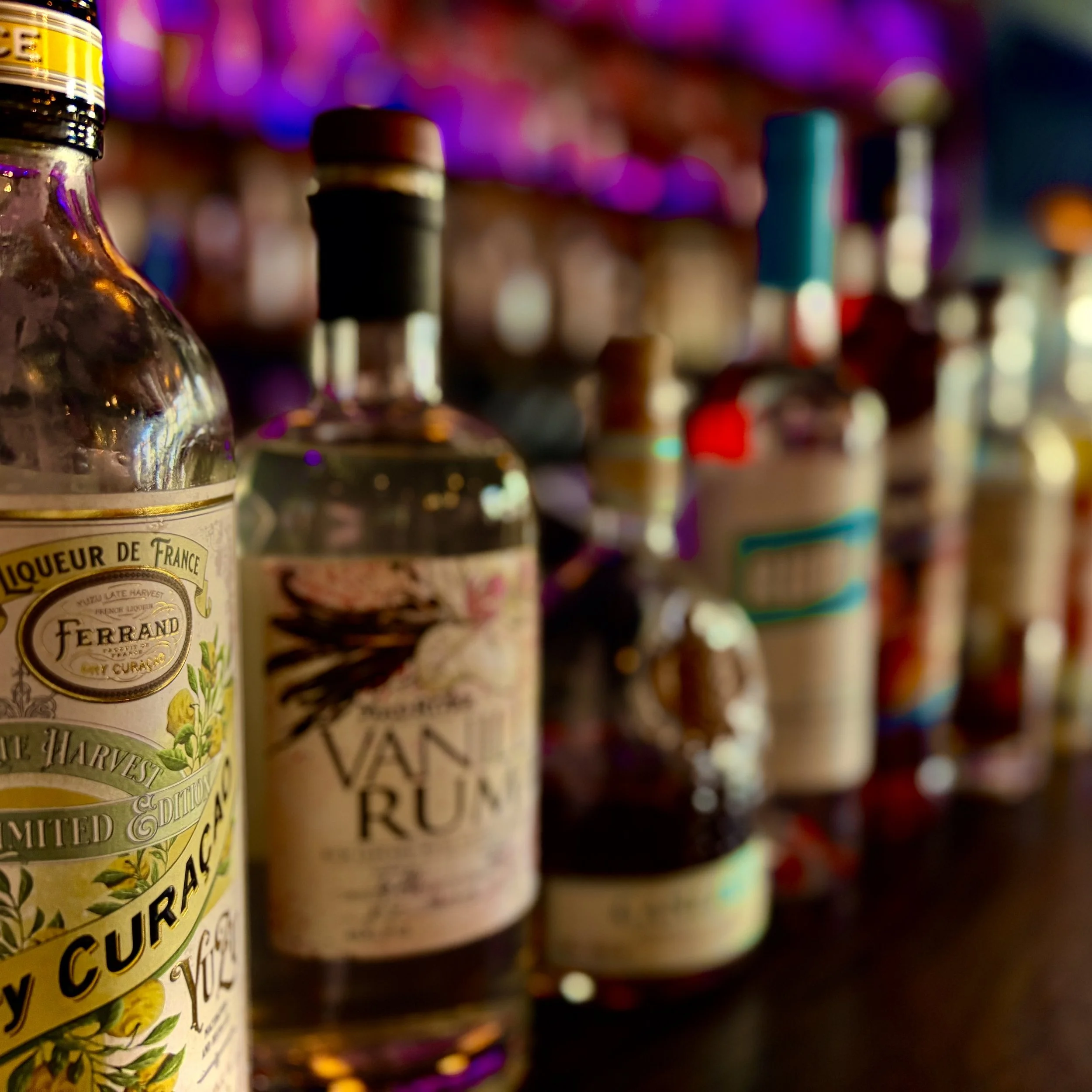 Close-up of various liquor bottles on a bar counter with a blurred purple-lit background.