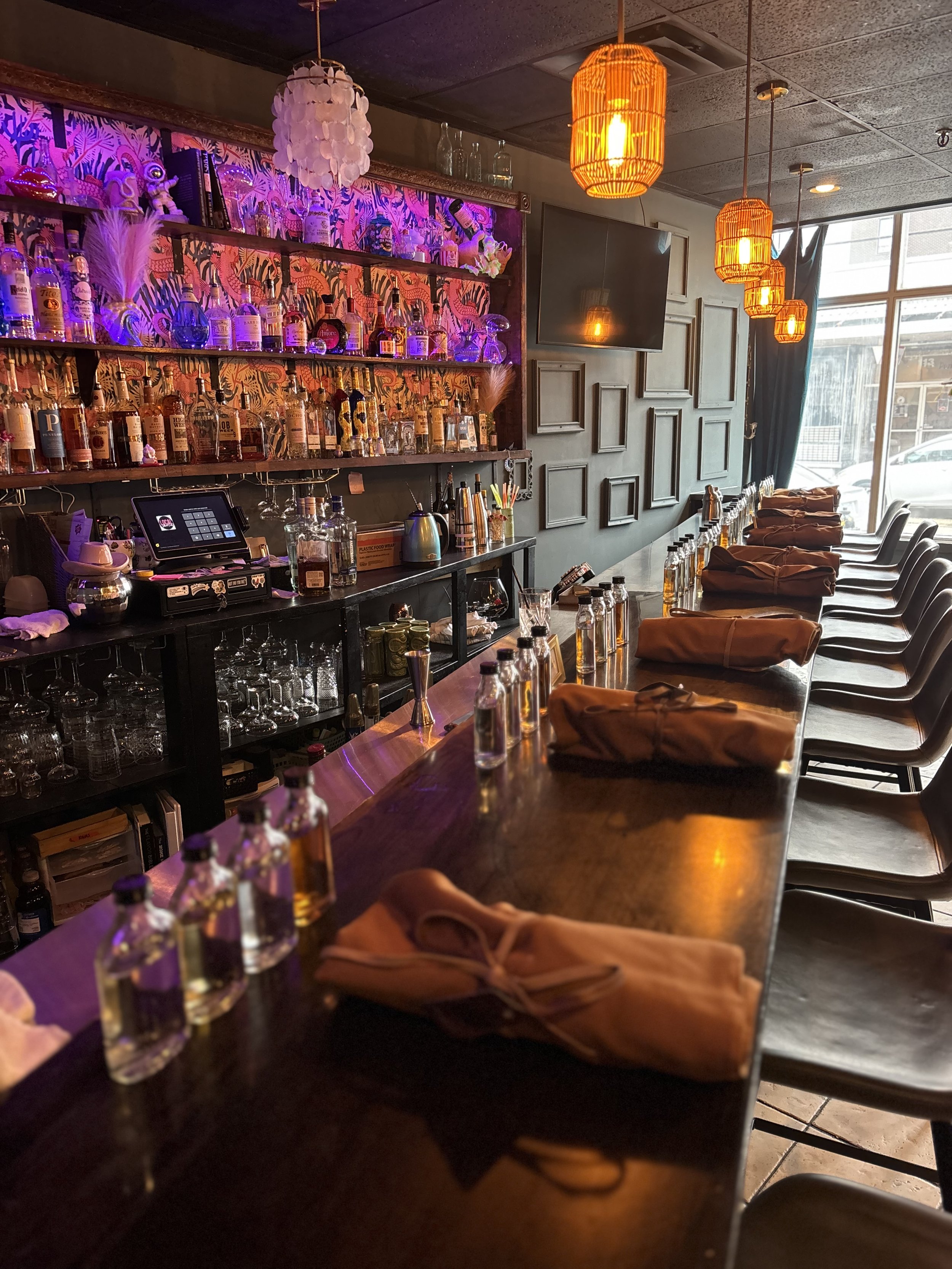 Bar interior with colorful lighting, bottles on shelves, counter seating with small bottles and rolled napkins, and pendant lights overhead. Shower, Celebration, Event, Private Rental, Party, Micro Wedding. 