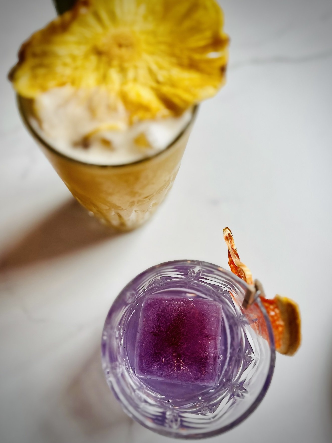 Two drinks on a table: one with a yellow flower garnish and another in a clear glass with a purple ice cube, garnished with dried fruit.