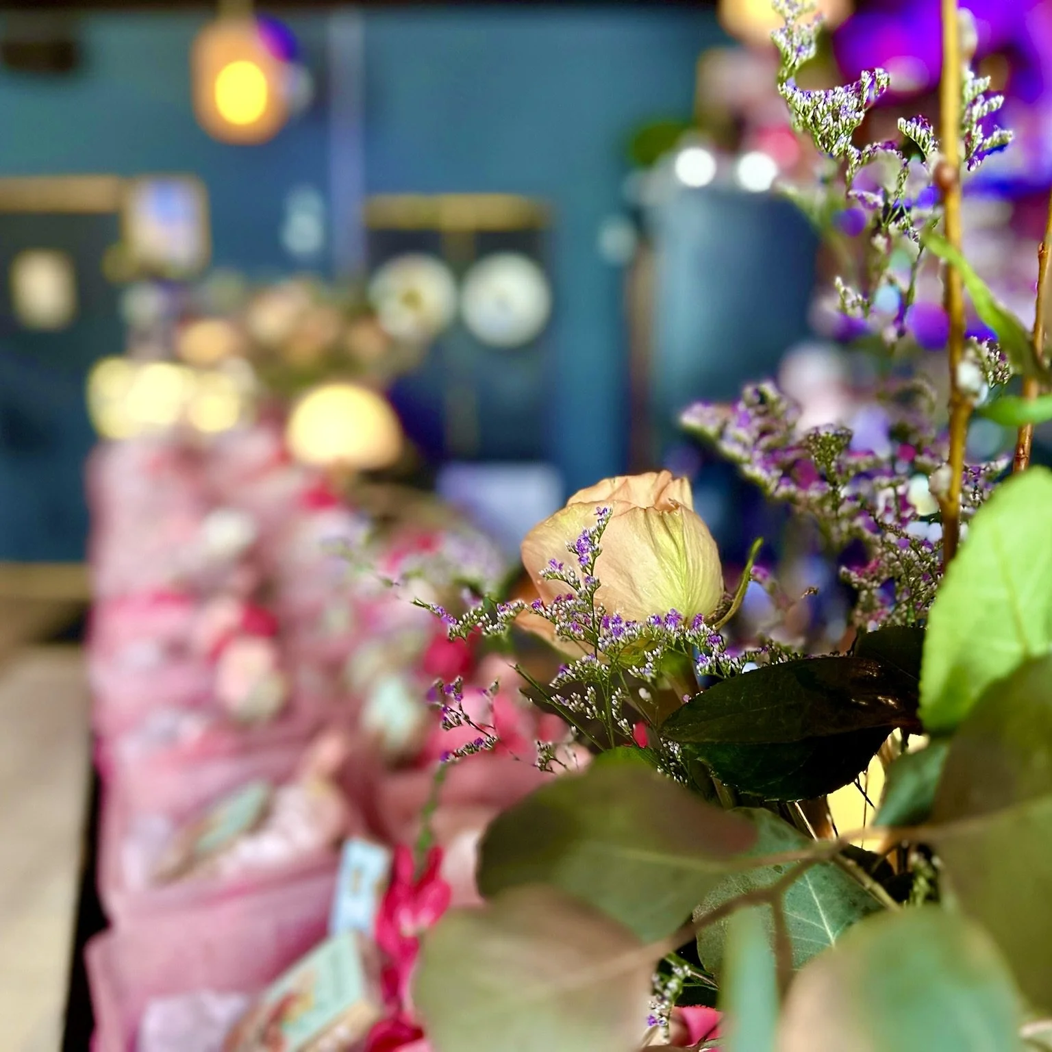 Close-up of a rose and purple flowers on a blurred background, possibly in a floral or gift shop setting.