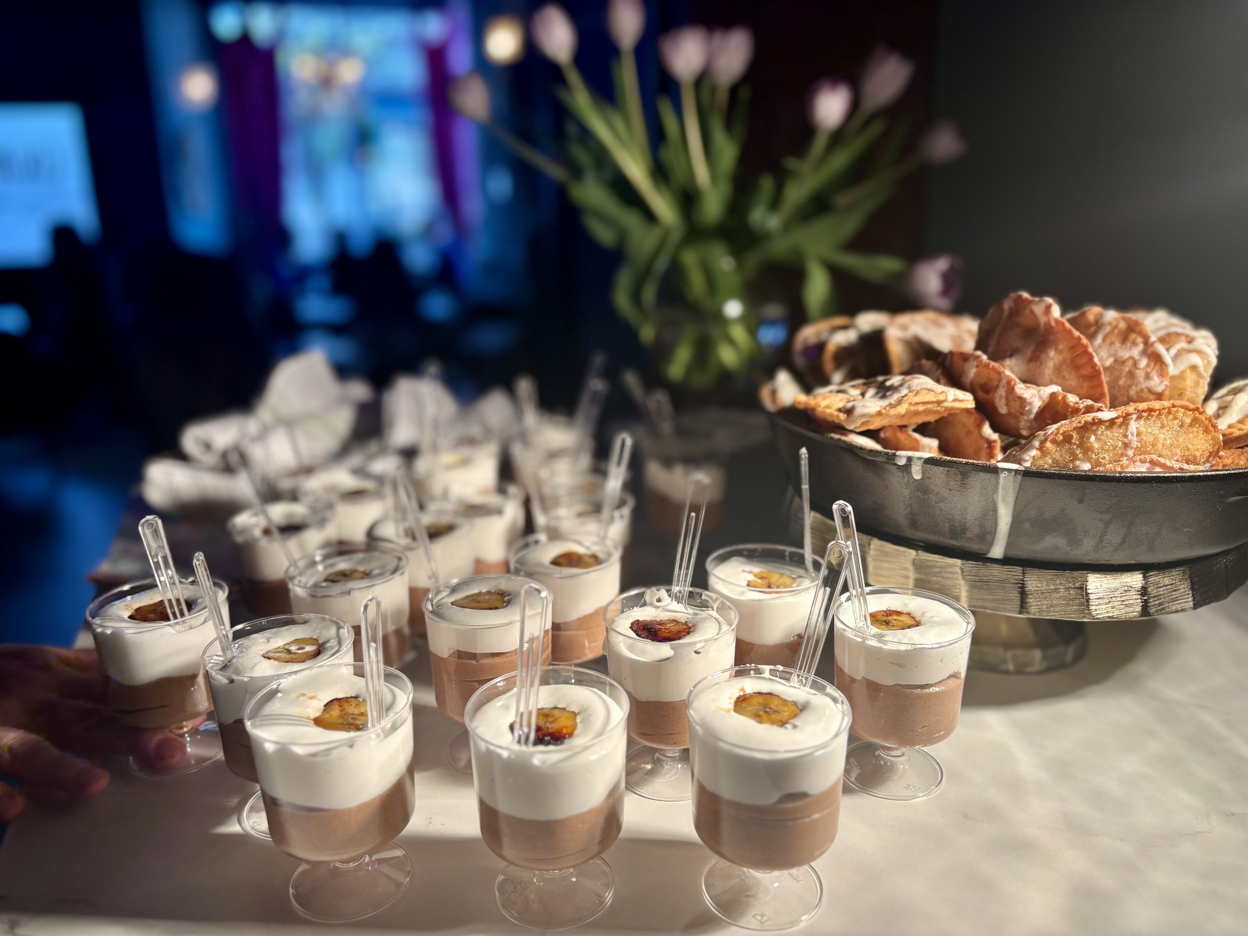 Dessert table with chocolate mousse cups topped with cream and banana slices, alongside a tray of glazed pastries, with blurred purple tulips in the background. Baby shower, wedding shower, wedding, celebration, private dining, private events, birthd