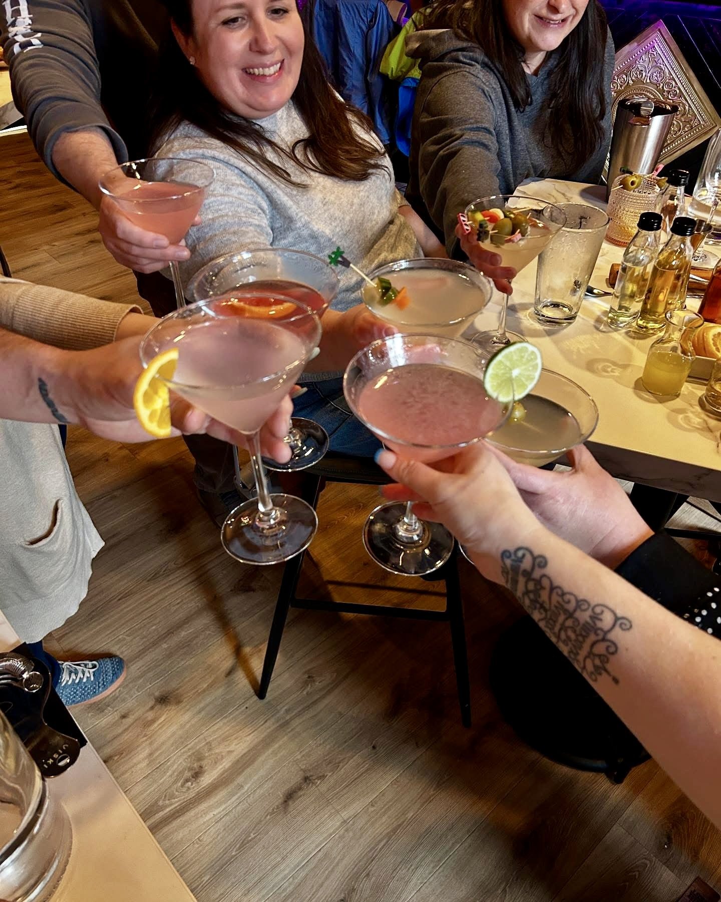 Group of people toasting with cocktails in a social setting, featuring various garnishes like lemon and lime slices. They are smiling and gathered around a table with drink bottles and glasses.