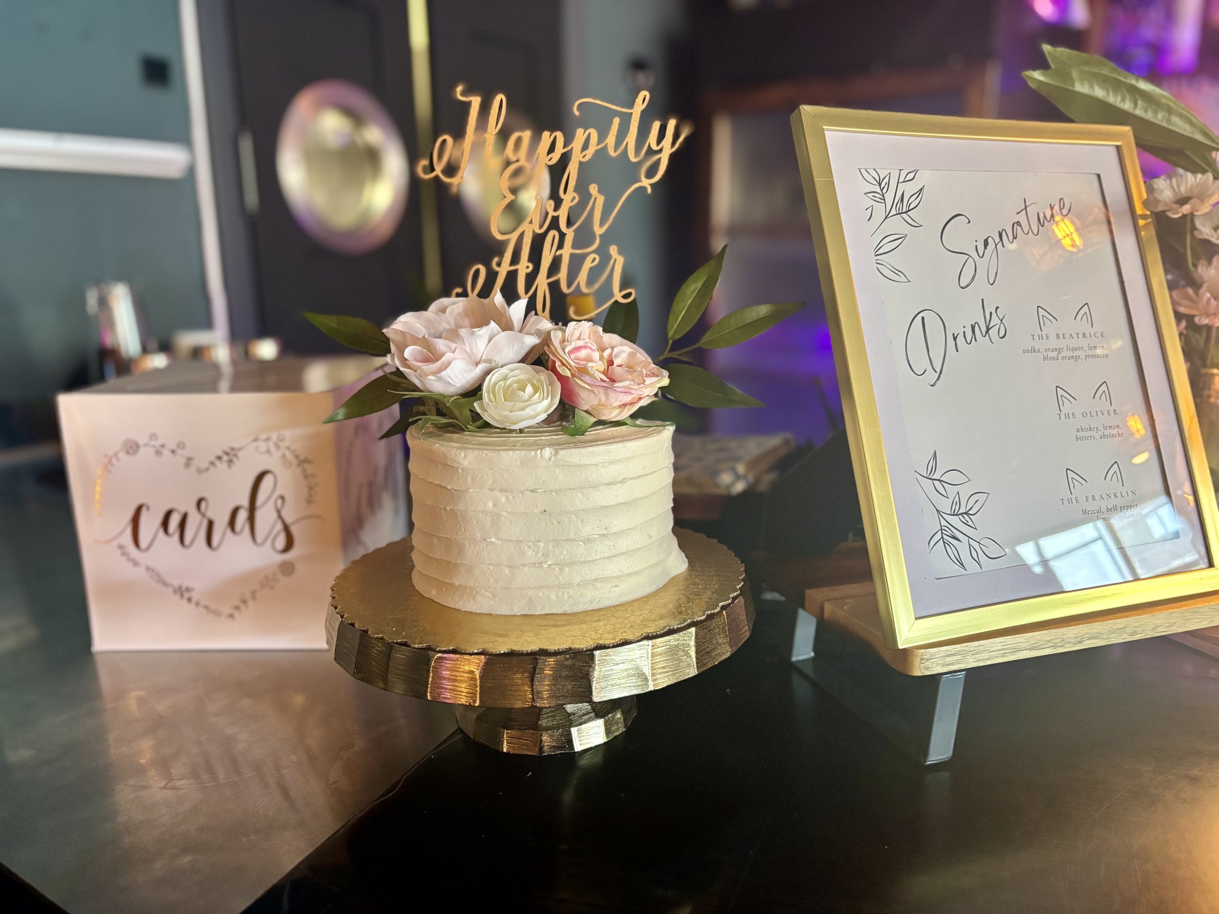 Wedding cake with "Happily Ever After" topper, surrounded by flowers, next to a "cards" box and a framed signature drinks menu on a table. Baby shower, wedding shower, wedding, celebration, private dining, private events, birthdays, custom menu. rest