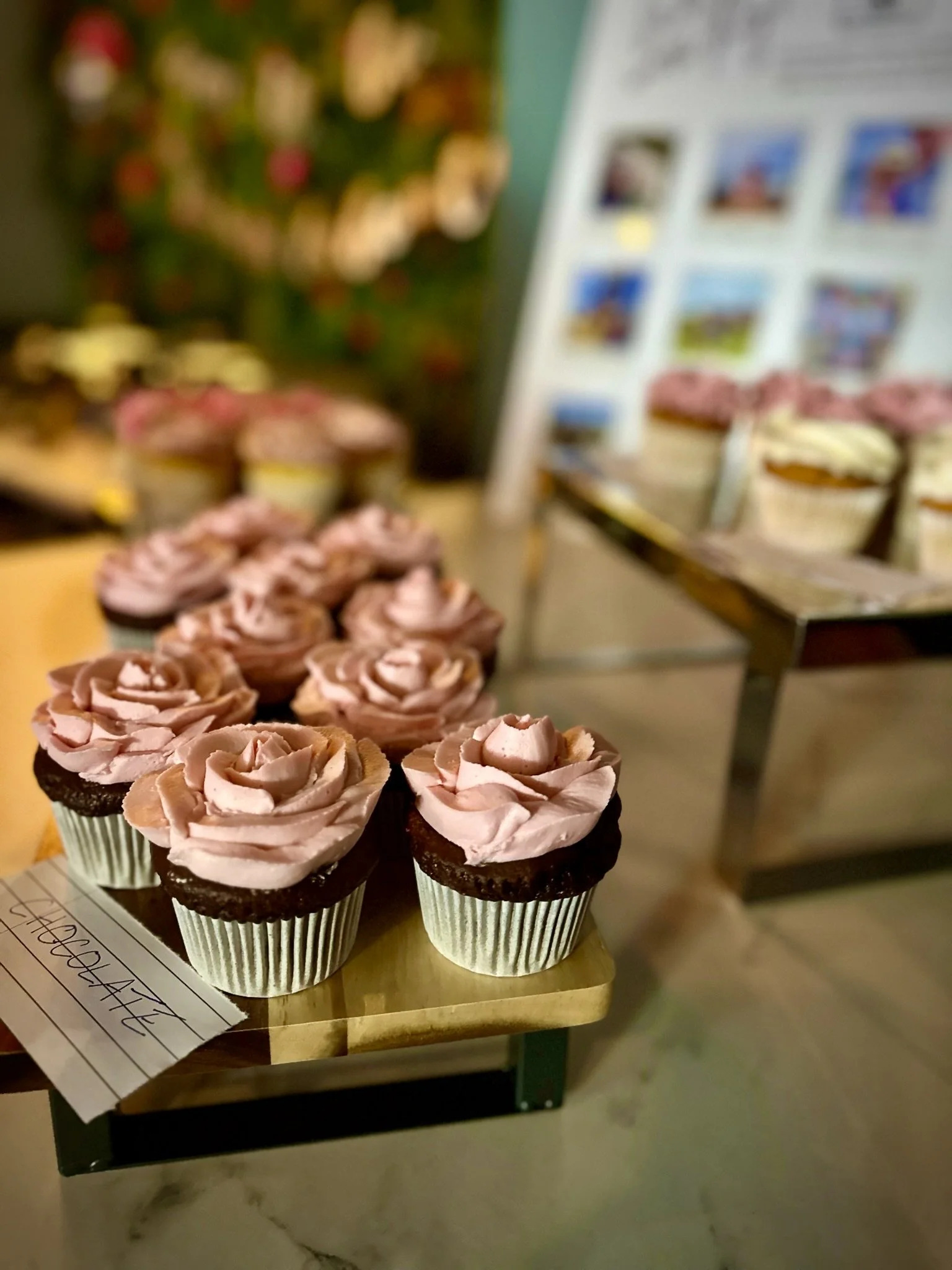 Cupcakes with pink rose-shaped frosting on a table, with more cupcakes in the background and colorful decorations on the wall.