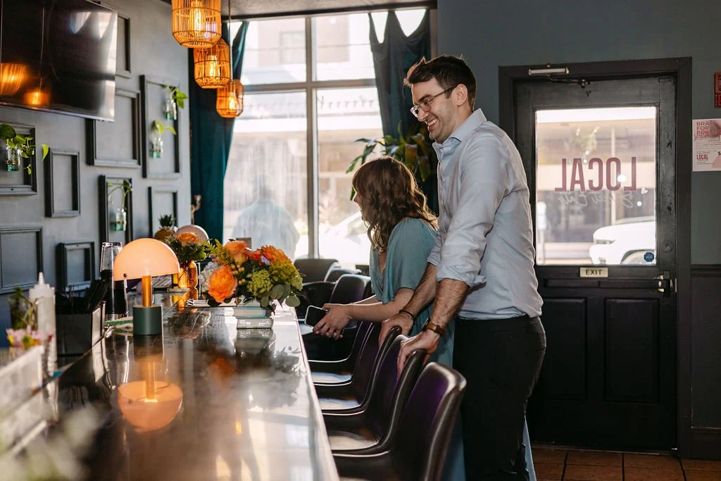A man and a woman standing at a bar counter inside a restaurant, smiling and looking at each other. The bar has a floral arrangement and a small lamp, with decorative hanging lights above. The restaurant has dark walls, large windows, and a sign on t
