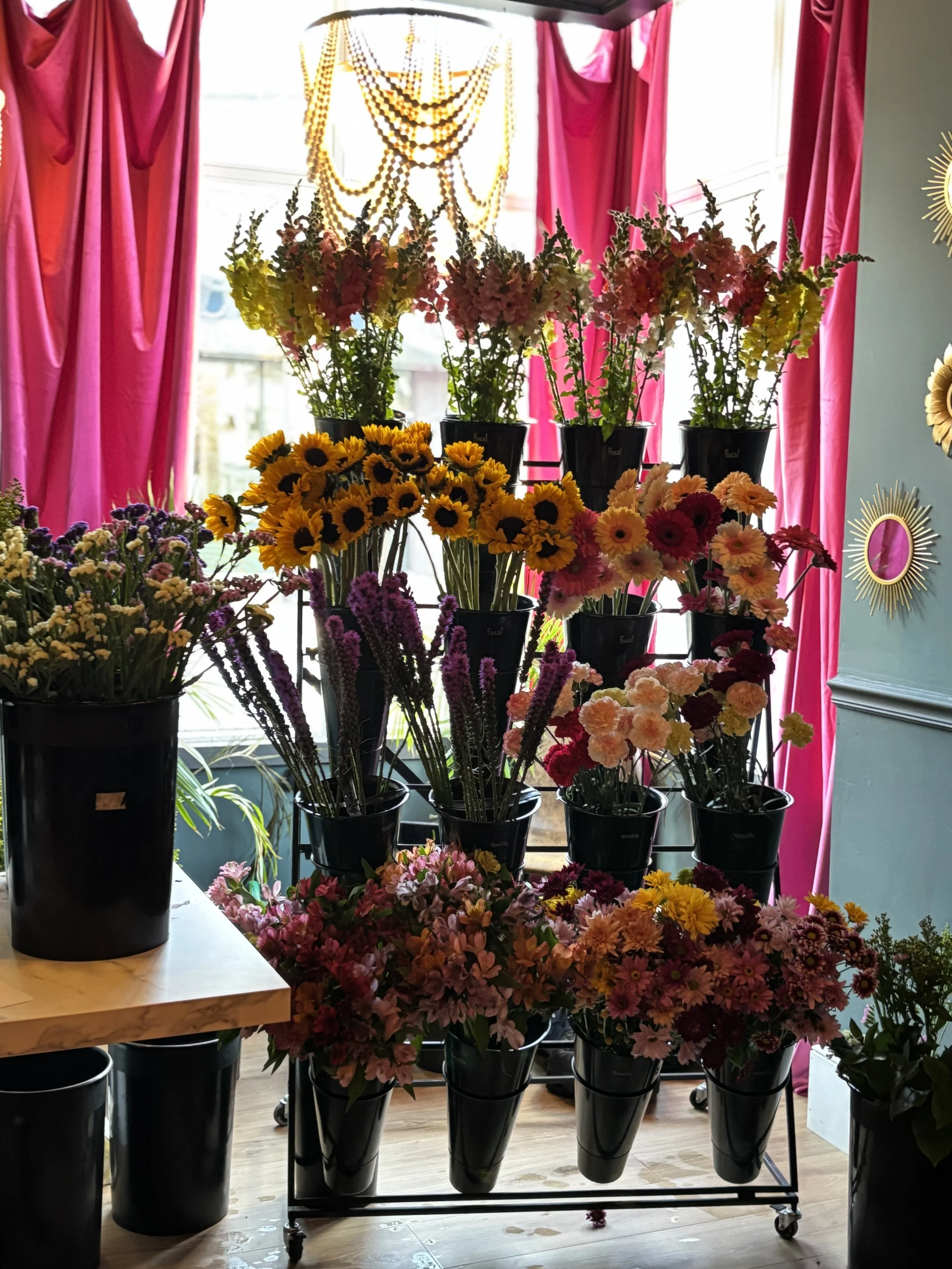 A display of various colorful flowers in black buckets, including sunflowers and snapdragons, inside a flower shop with pink curtains and a beaded chandelier.