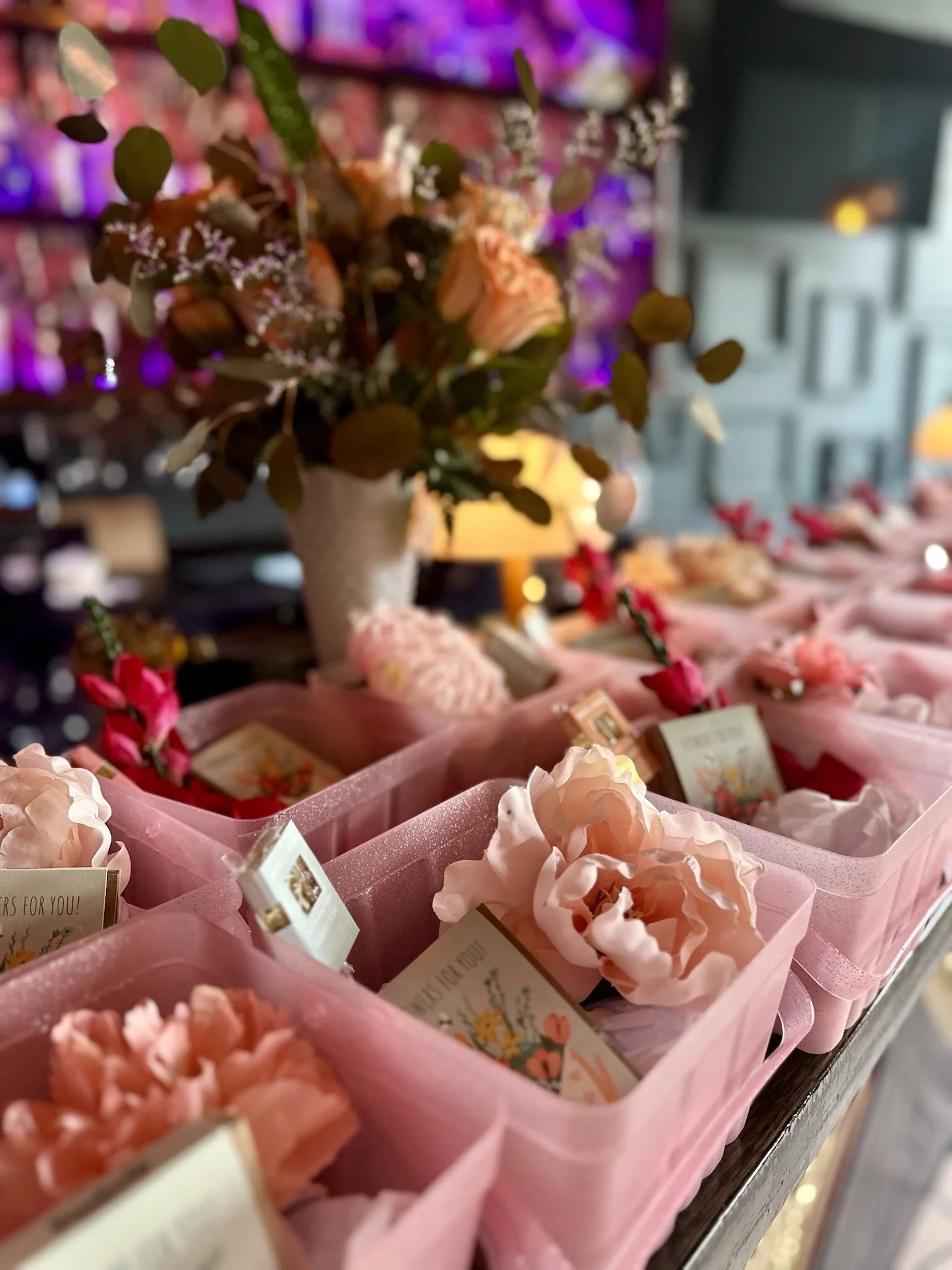 Pink baskets containing flowers and small cards on a table, with a floral arrangement in the background.