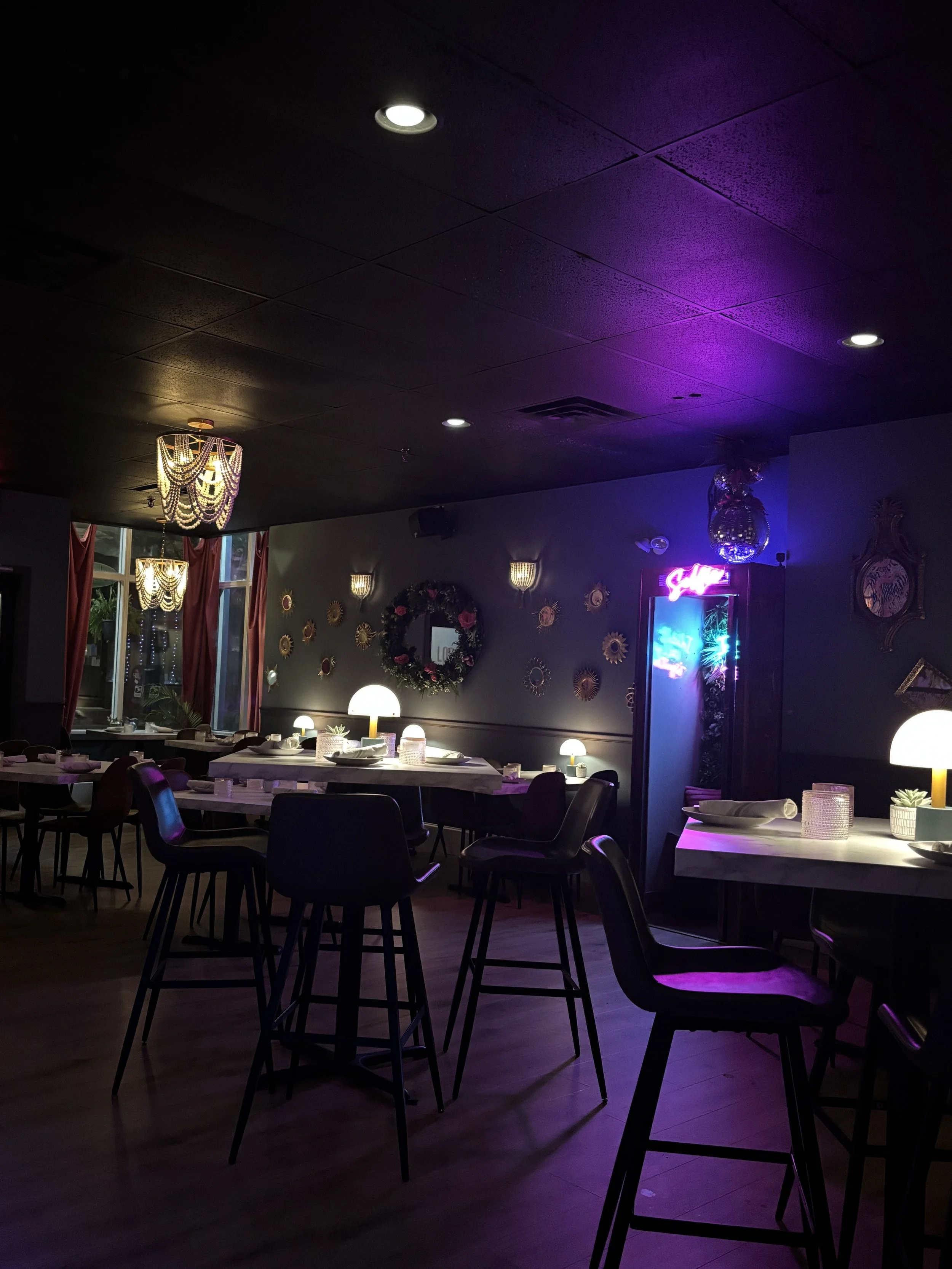 Dimly lit restaurant with black ceiling, illuminated tables, bar stools, chandeliers, wall decor, and neon sign.