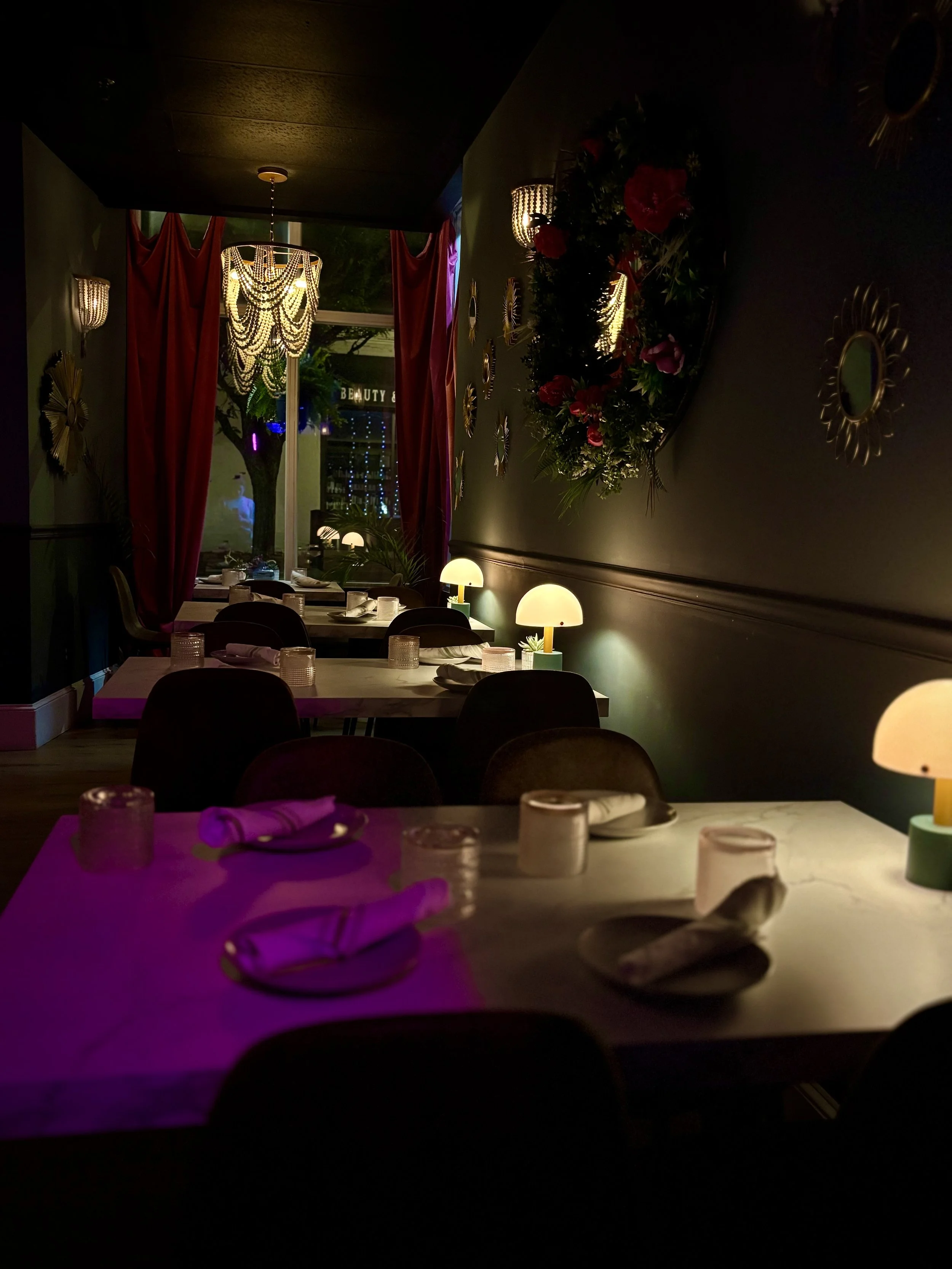Dimly lit restaurant with tables set with napkins, glasses, and plates, decorated with floral arrangements, lamps, and a chandelier, featuring a window with red curtains.