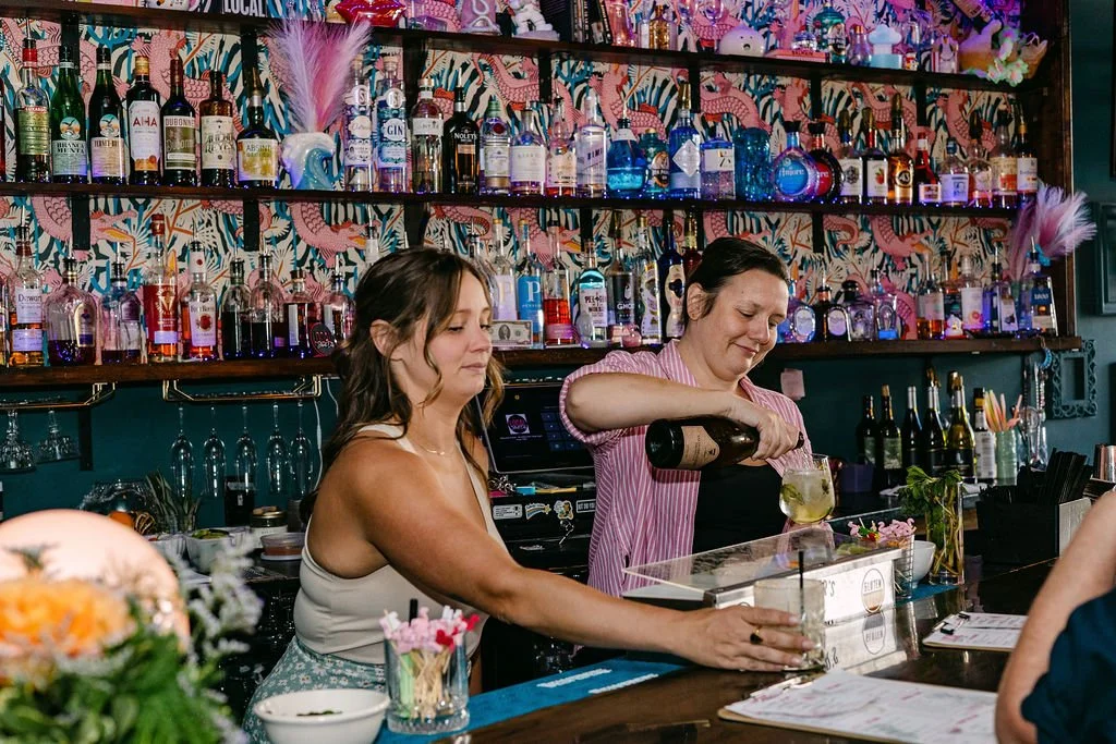 A woman two women behind a bar, with one pouring a drink into a glass, decorated with colorful bottles and a vibrant pink and botanical wallpaper background.