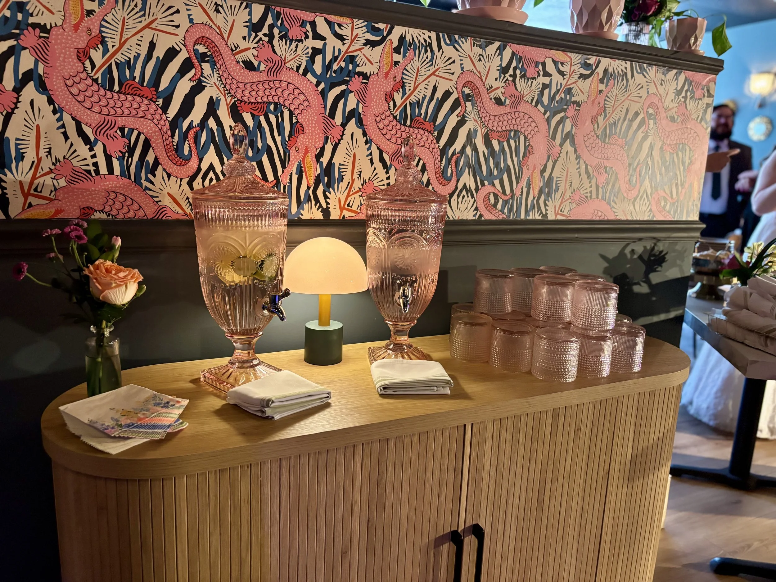 Glass beverage dispensers and stacked glasses on a wooden counter with colorful dragon print wallpaper in the background, alongside flowers and napkins. Micro Wedding 