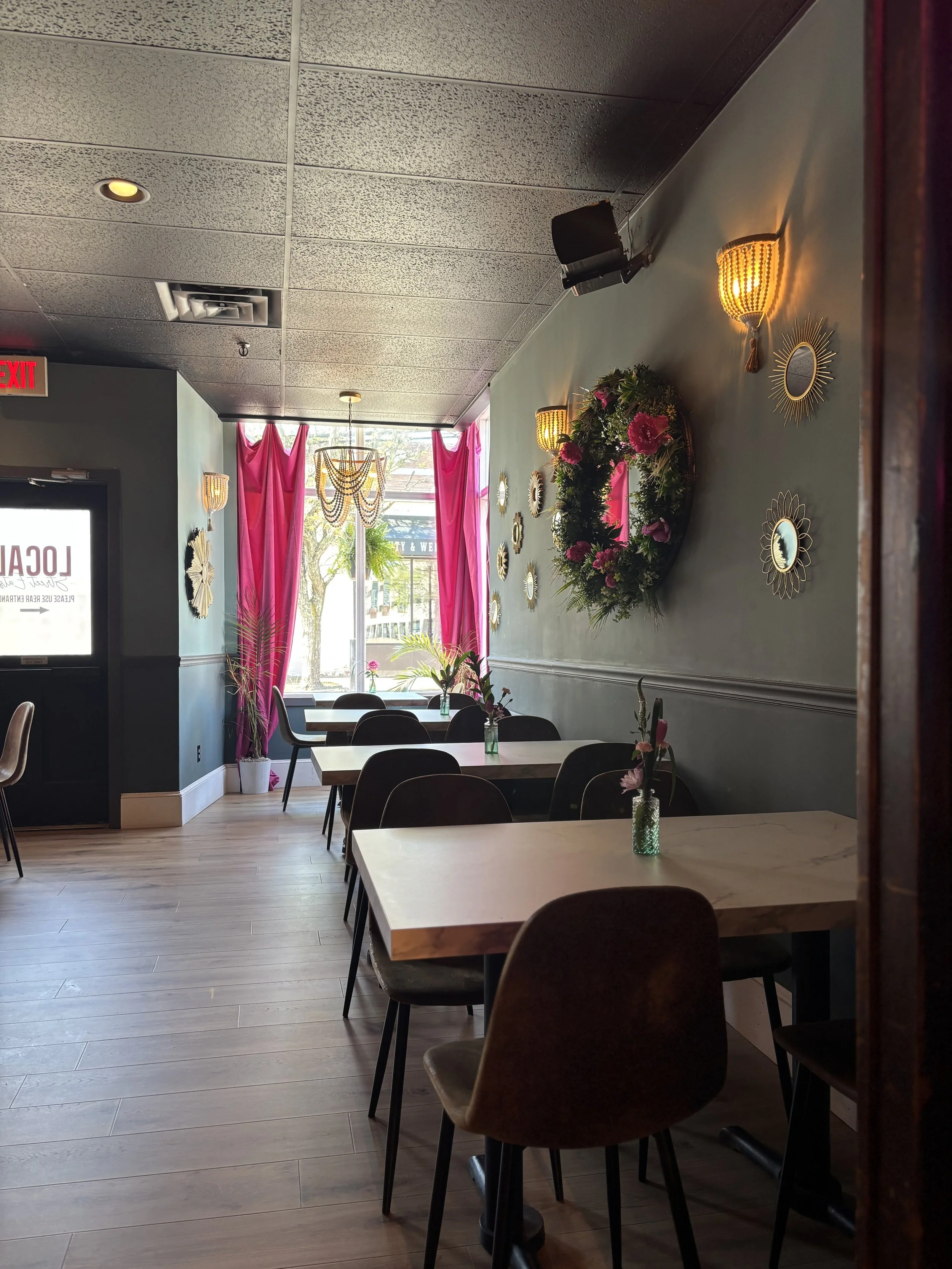 Interior of a cozy cafe with tables and chairs, a wall decorated with gold-framed mirrors and a floral wreath, pendant lighting, and pink curtains on the window. Private Events Rentals Best of Nashua NH Best Street Food Smash Burgers Downtown Nashua