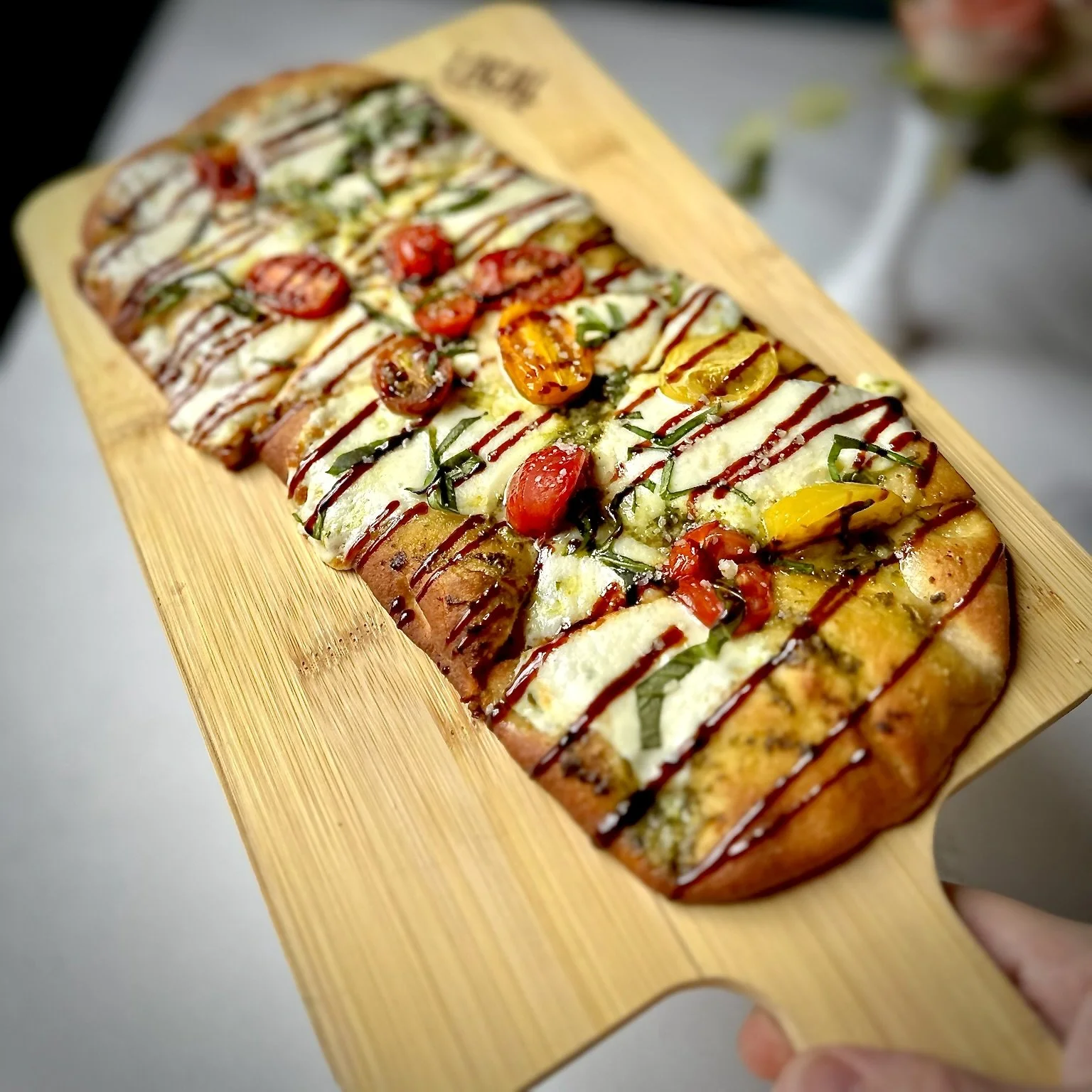 Flatbread pizza on a wooden board, topped with cherry tomatoes, mozzarella cheese, basil, and drizzled with balsamic glaze.