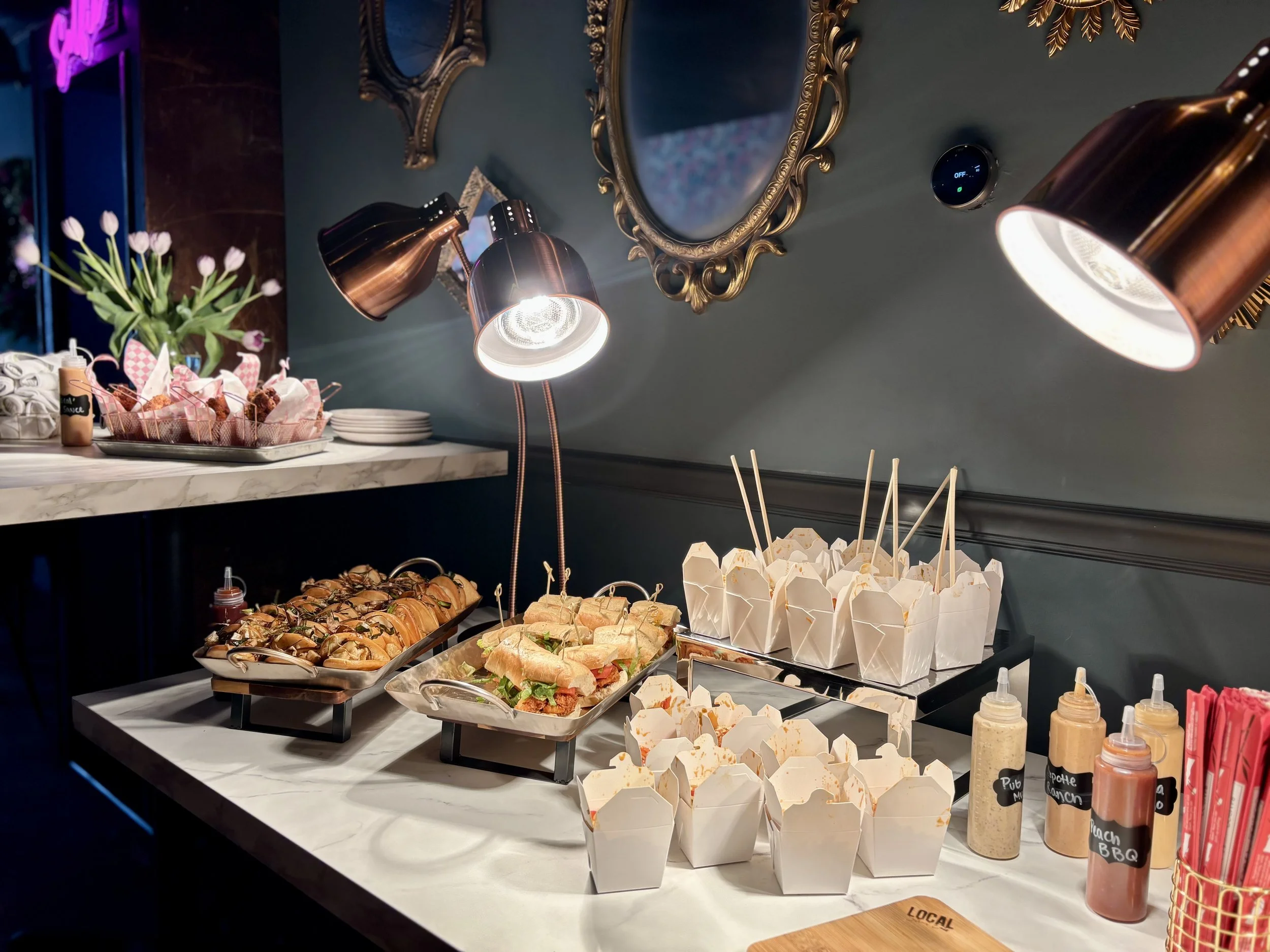 Buffet with sandwiches, boxed meals, and sauce bottles under warm lighting, decorated with flowers and mirrors.