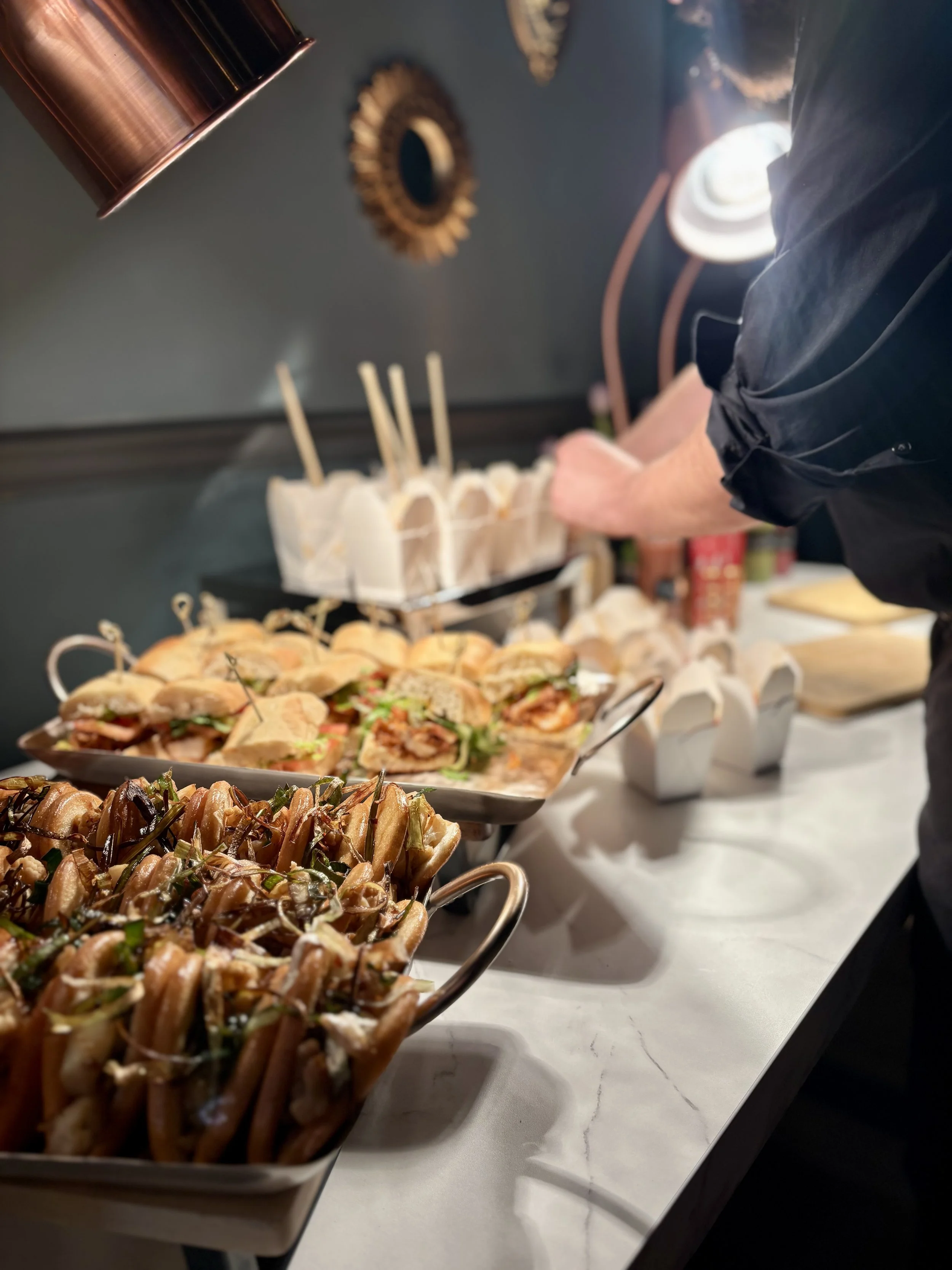 Person arranging gourmet sliders and hot dogs on a counter under a heat lamp, surrounded by packaging boxes. Shower, Celebration, Event, Private Rental, Party, Micro Wedding. 