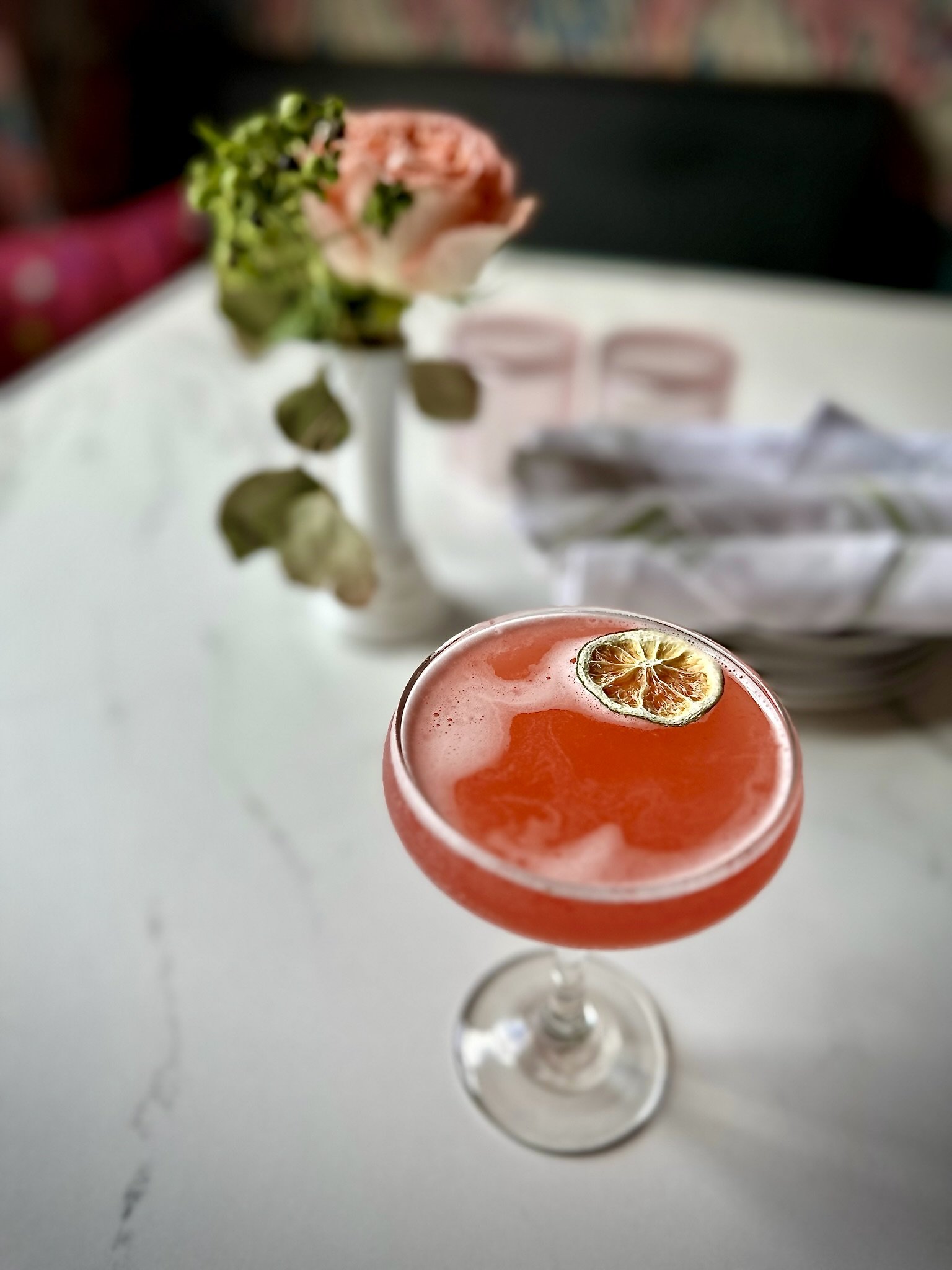 Pink cocktail with a dried lime garnish in a coupe glass on a white marble table, with a blurred background of a rose in a vase and folded napkins.