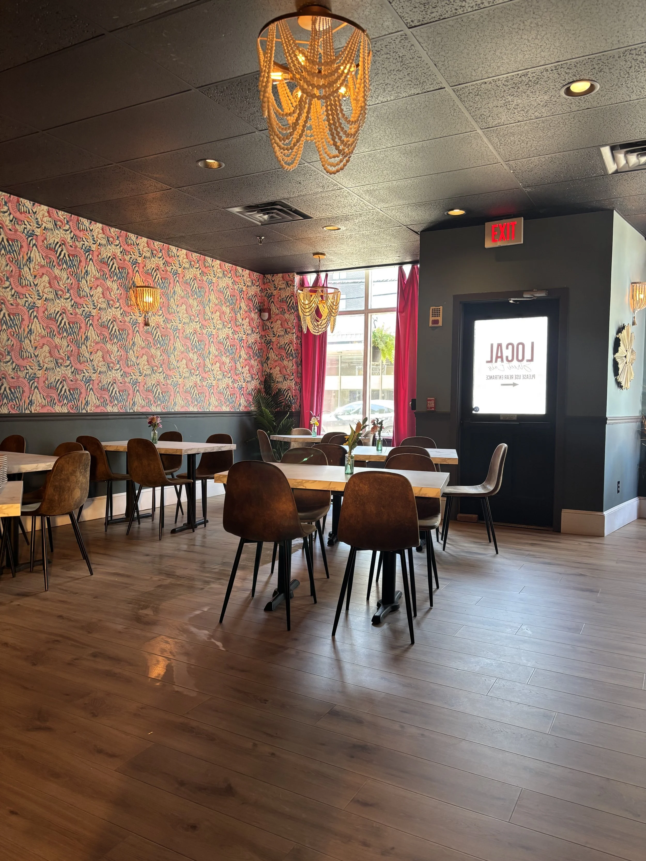 Chic restaurant interior with modern decor, featuring pendant lights, floral wallpaper, and wooden flooring, with several tables and chairs arranged for dining.