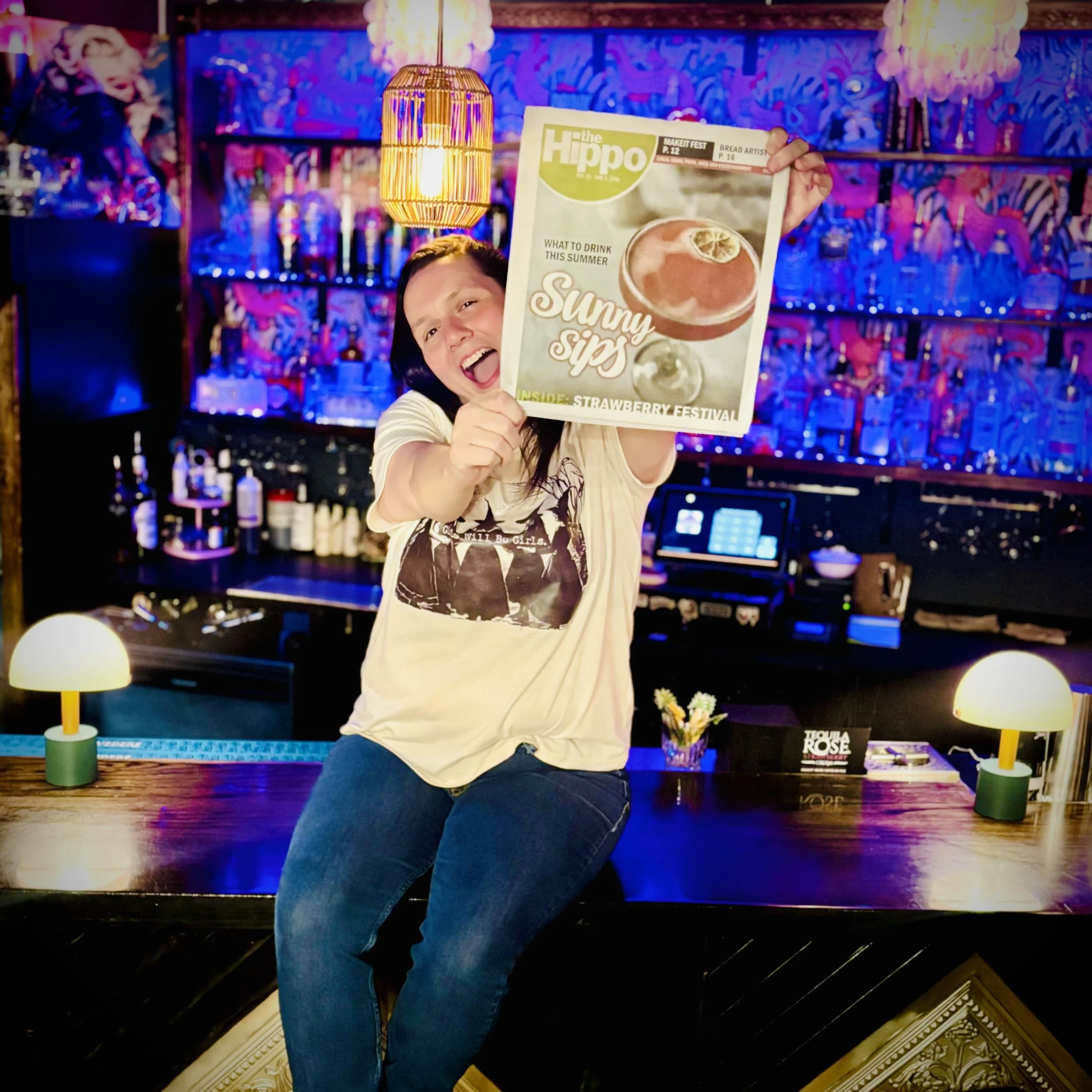 A woman sitting on a bar counter holding a newspaper with a cheerful expression. The background is decorated with colorful, vibrant wall art and warm lighting, including two small lamps on the bar surface.