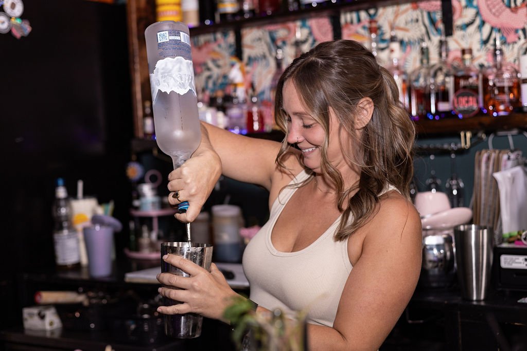 A woman smiling and pouring a clear liquid from a bottle into a glass behind a bar or countertop in a lively setting.