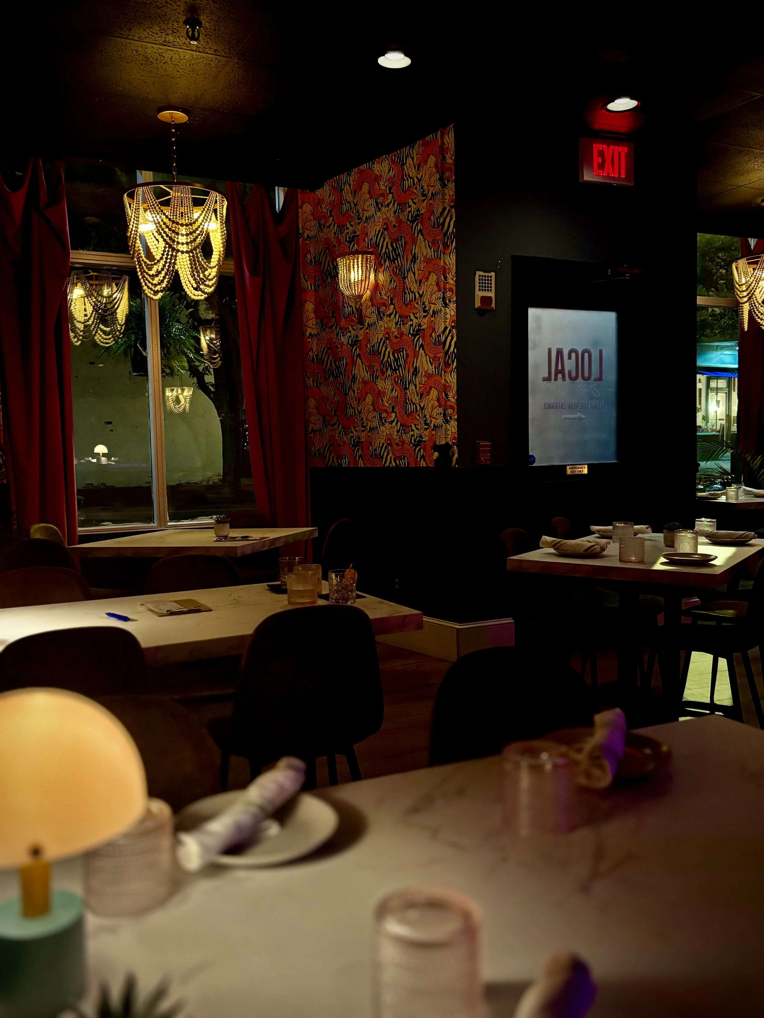Interior of a dimly lit restaurant with windows, red curtains, chandeliers, and various tables set with napkins, glasses, and plates.