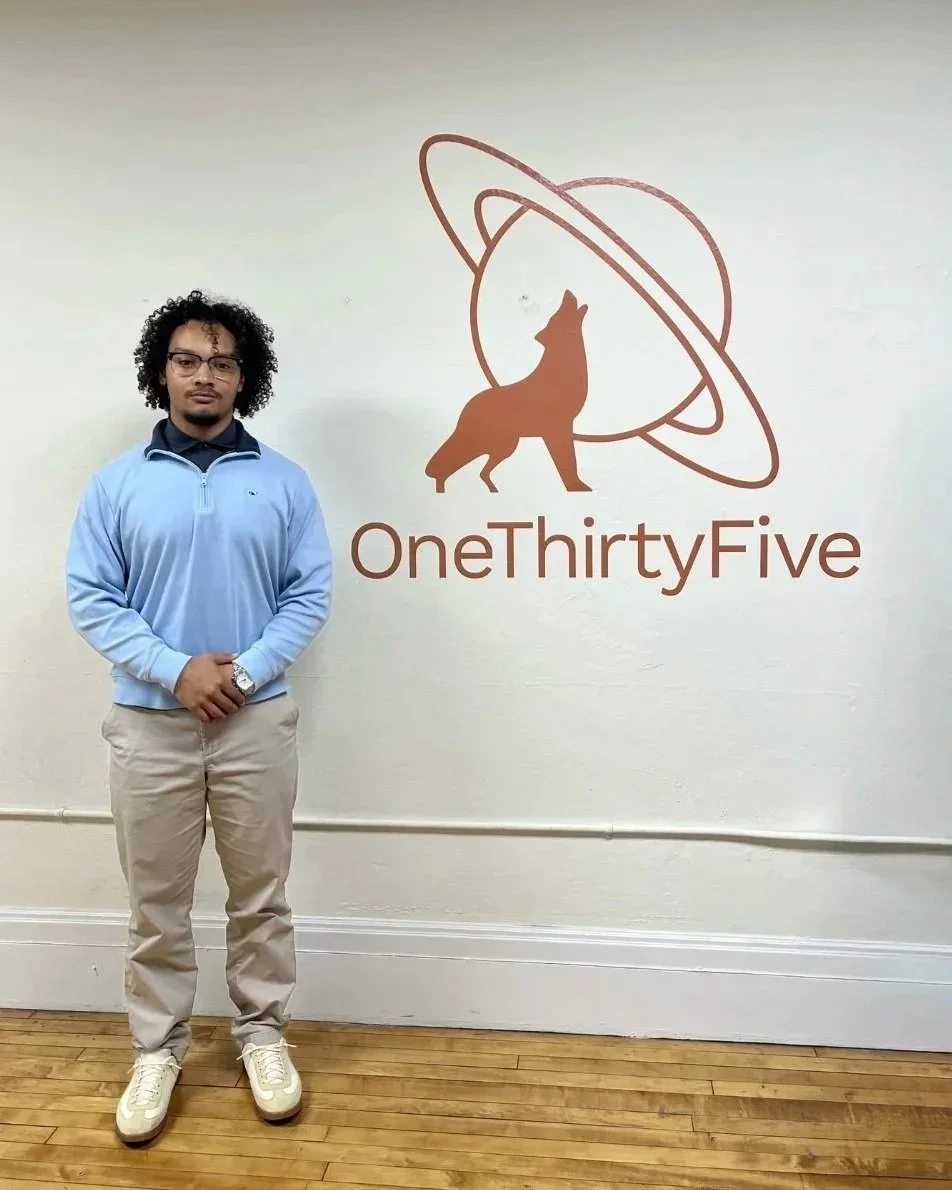 A young man with curly hair and glasses standing in front of a wall with a logo of a wolf and a planet with rings, and the text 'OneThirtyFive'. He is wearing a light blue jacket, beige pants, white sneakers, and a watch.