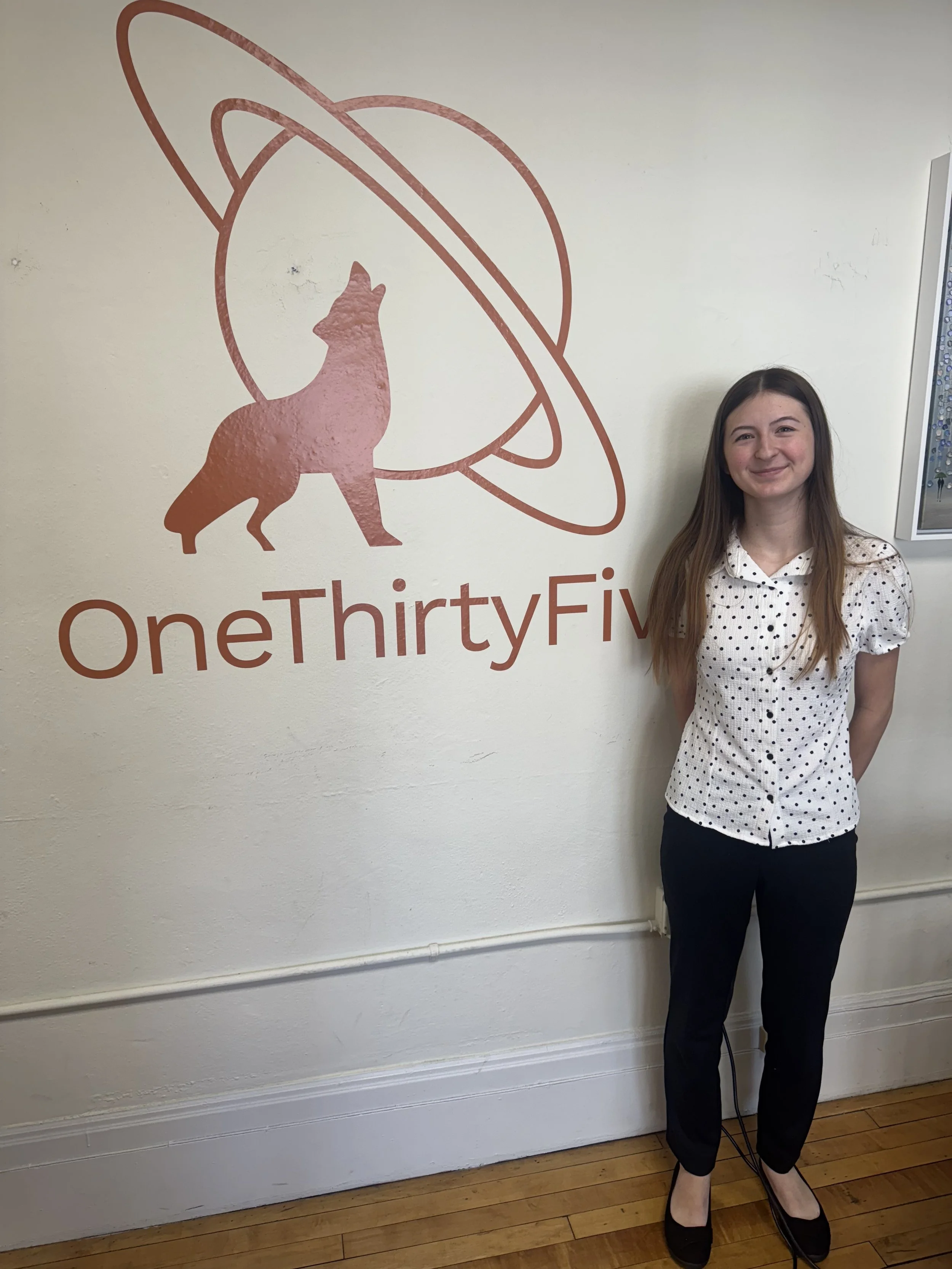 A young woman with long brown hair smiling, standing indoors against a light-colored wall with a logo that says 'OneThirtyFive' and features a pink silhouette of a howling dog with stylized orbits around its head.