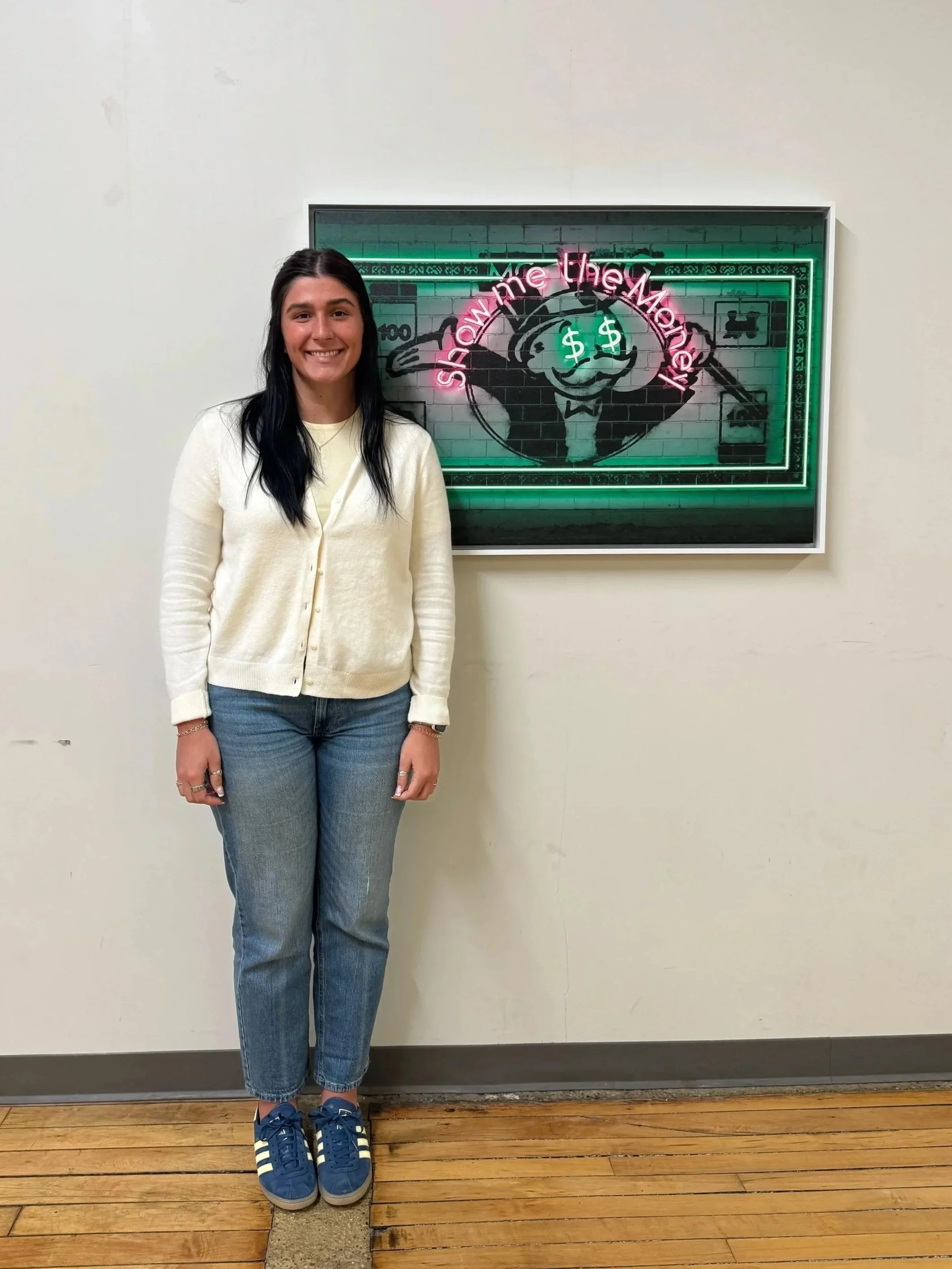 A young woman standing in front of a white wall and a neon sign that reads 'Show Me the Money' with a cartoon character with dollar signs in its eyes, wearing a tuxedo, in a frame.