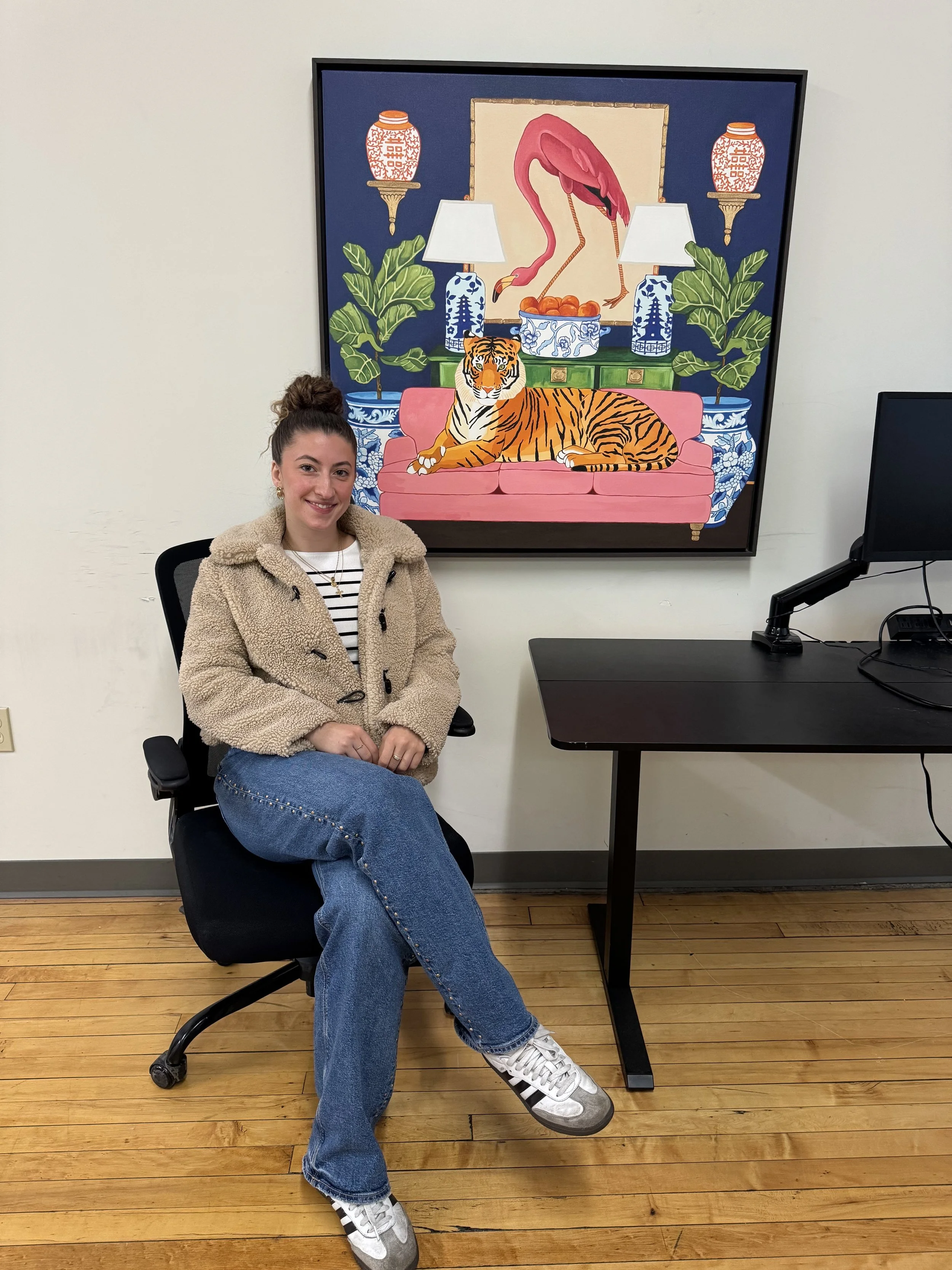 A young woman with brown hair in a bun, wearing a beige fuzzy coat, striped shirt, and blue jeans, sitting in a black office chair in front of a white wall with a colorful painting of a pink flamingo, green plants, a tiger on a pink sofa, and decorative lamps.