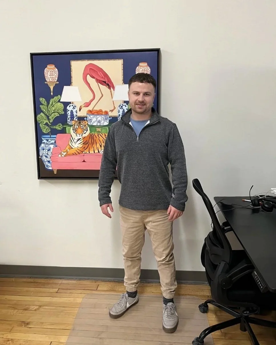 A man standing in an office space with a colorful artwork of a pink flamingo, tiger, and decorative vases behind him. He is wearing a gray zip-up jacket, khaki pants, and white sneakers.