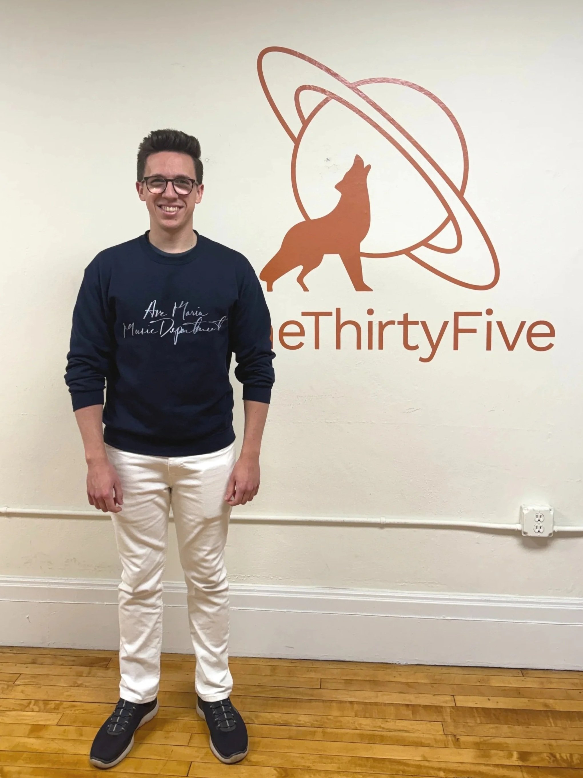 A young man with glasses, wearing a navy blue sweatshirt and white pants, standing in front of a wall with the logo and name 'eThirtyFive'. The logo features a fox, a planet with rings, and a stylized font.