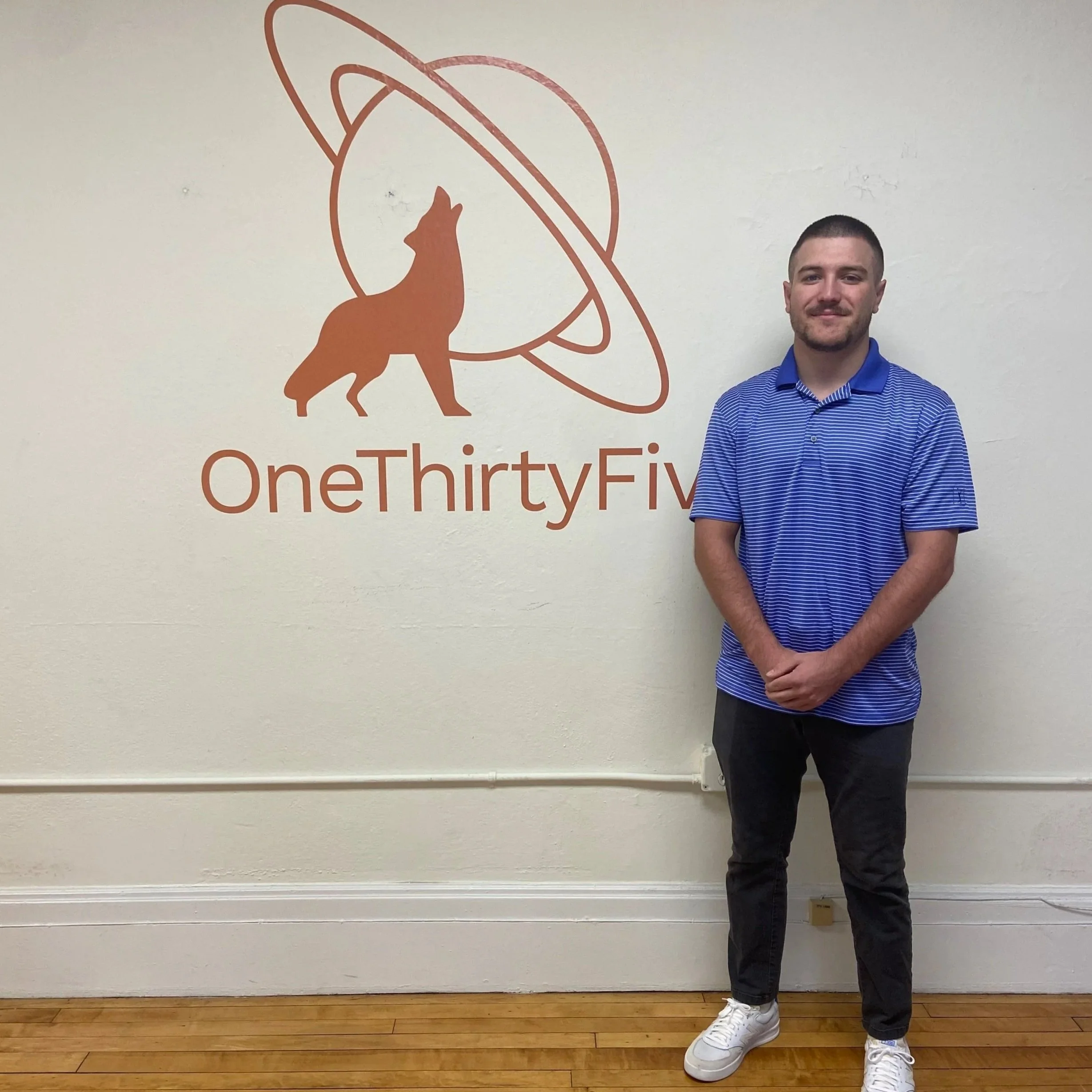 A man in a blue striped polo shirt and black pants standing in front of a wall with a logo of a dog and a planet, with the text "OneThirtyFive" below it.