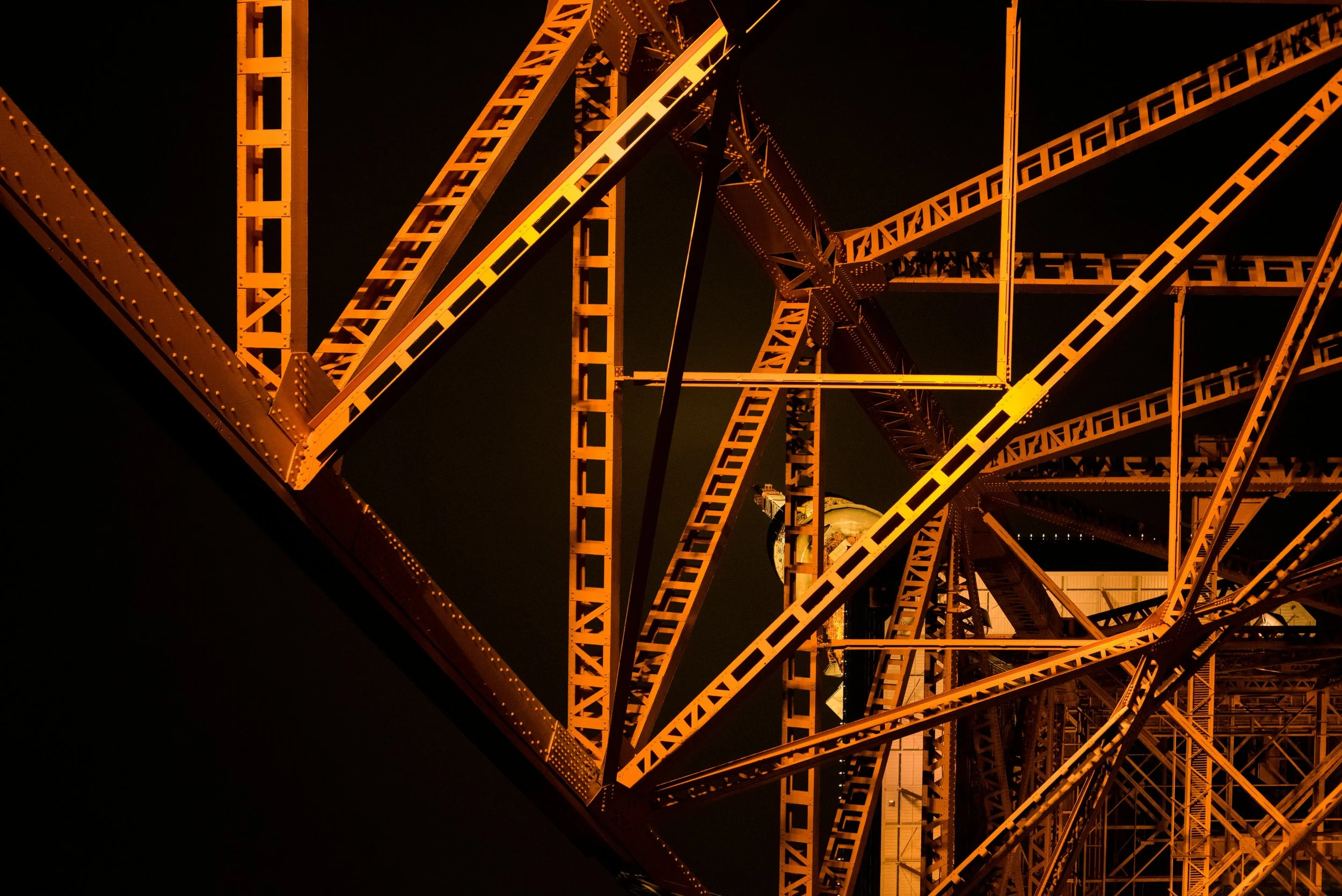 Close-up shot of the intricate steel framework of a bridge at night, illuminated by orange lighting, highlighting the structural details and riveted connections.
