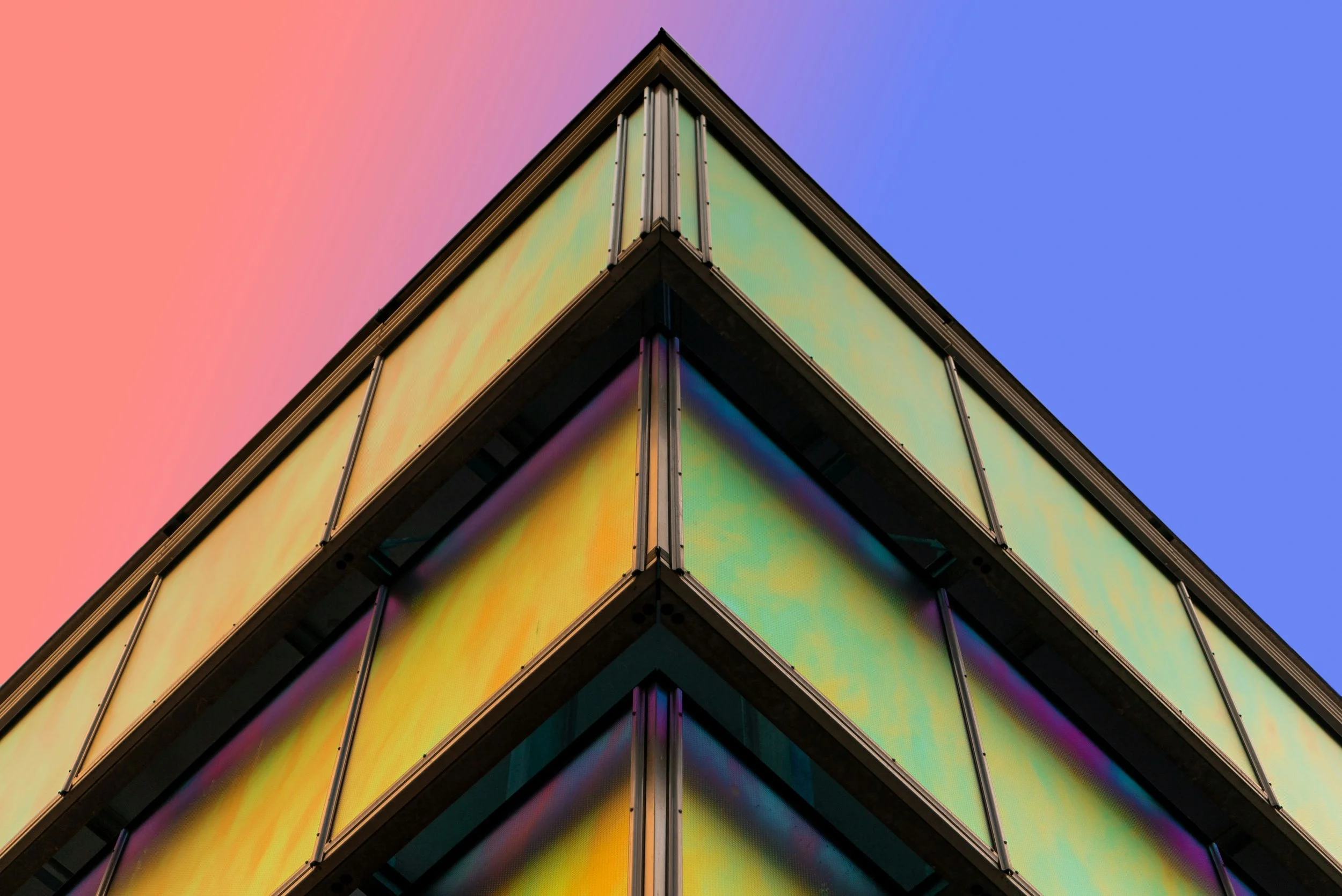 Close-up view of a modern building corner with reflective glass panels displaying a gradient of colors, set against a pastel pink and blue sky
