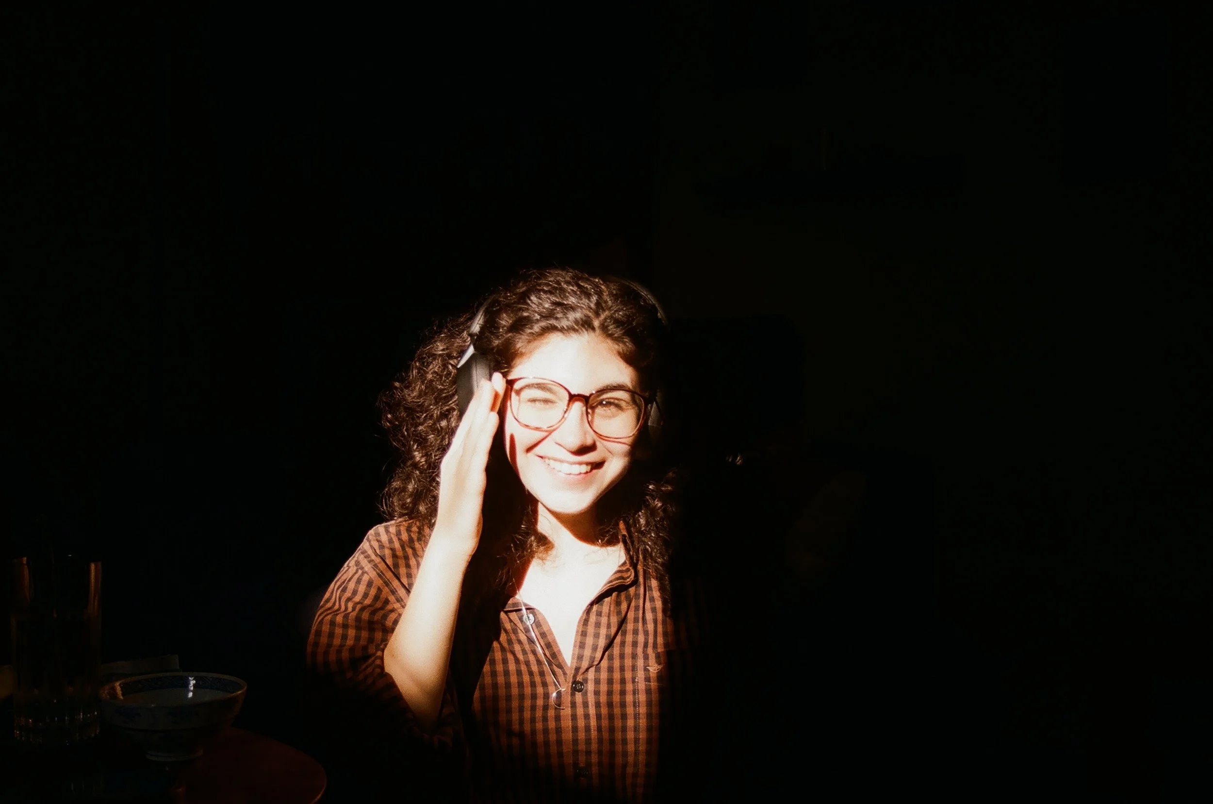 high contrast photograph of Emma smiling and squinting into the sun. She holds her hand up to shield her eyes. The surroundings are black.