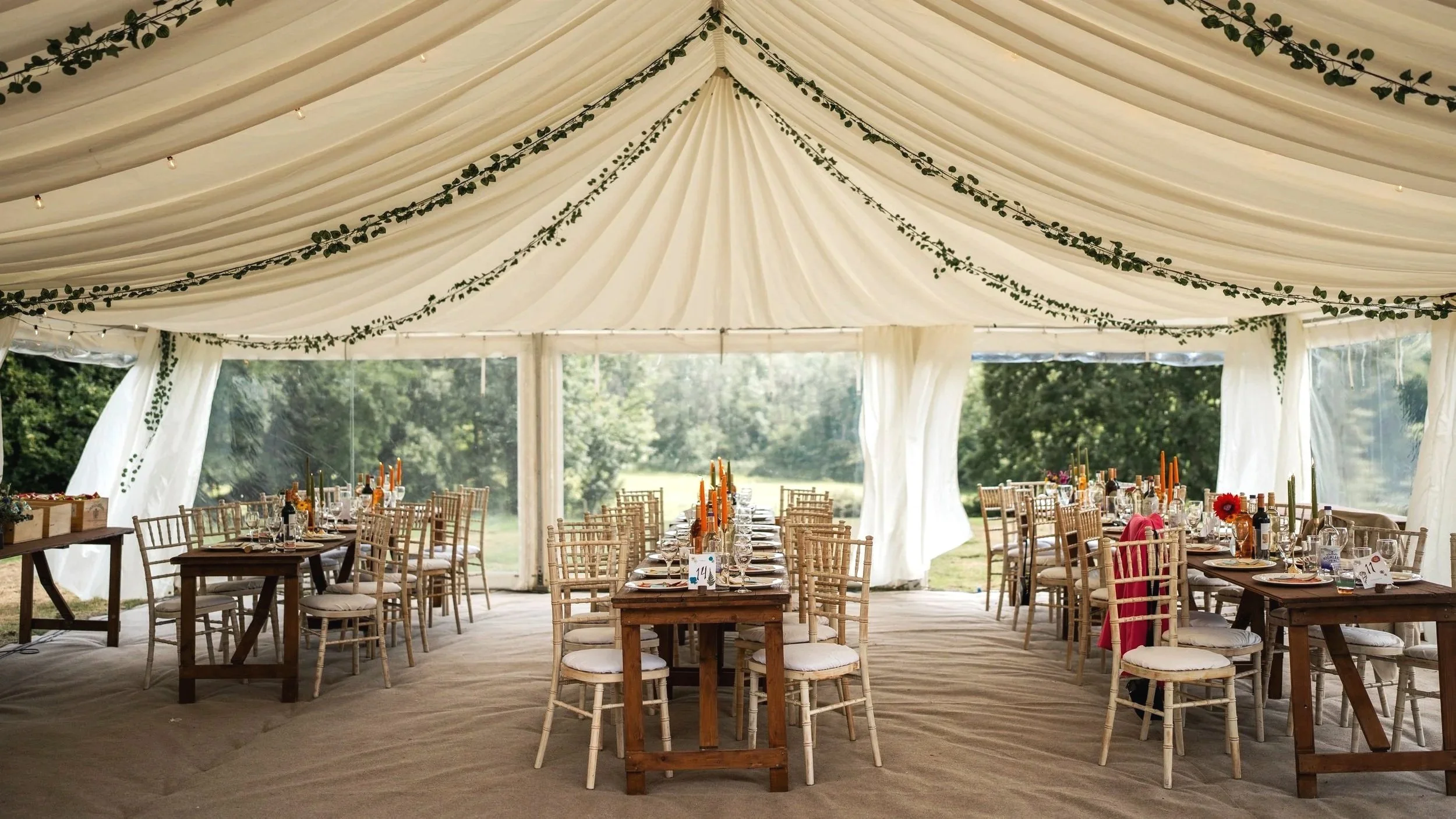 Wedding reception tent with decorated tables, chairs, and greenery, outdoors with trees visible through the tent walls.