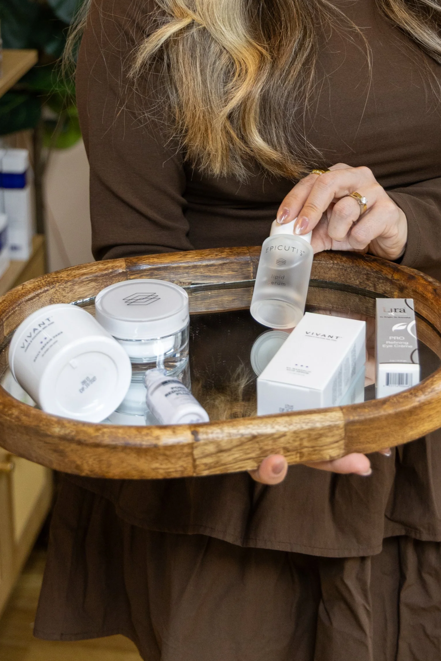 A woman holding a wooden tray with skincare products, including jars, bottles, and boxes, on a mirrored surface.