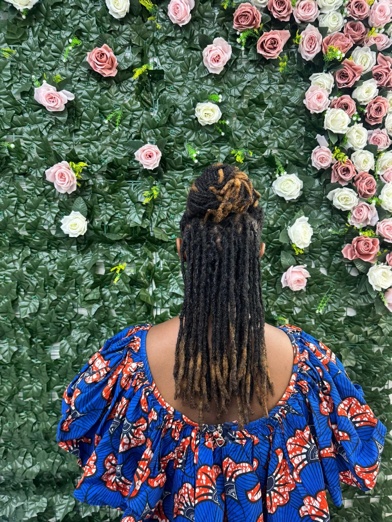 A woman with long dreadlocks wearing a colorful blue and red patterned dress, standing in front of a wall decorated with pink and white roses and green leaves.