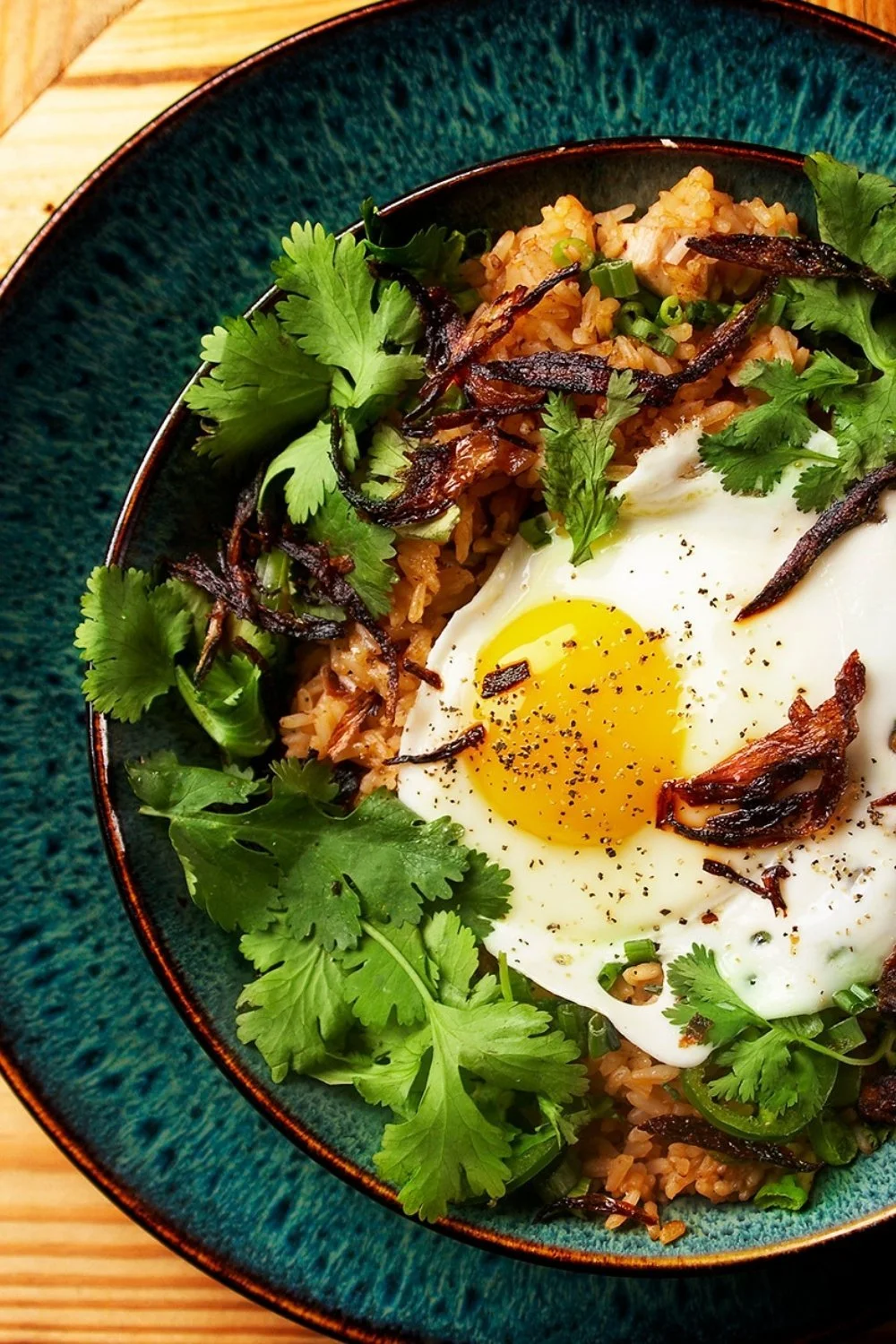A plate of fried rice topped with a sunny-side-up egg, garnished with fresh cilantro and crispy fried onions.