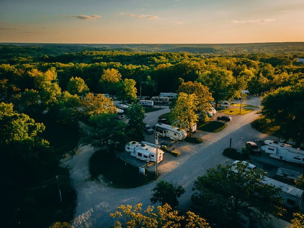 An aerial view of a campground with numerous RVs and campers parked among green trees during sunset.