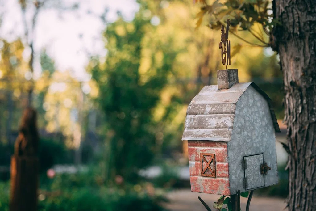 A decorative metal mailbox designed to look like a small house, mounted on a tree trunk in a garden or yard, with greenery and trees in the background.