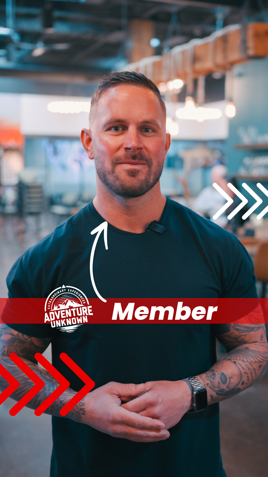 A man with tattoos on his arms and short hair standing inside a modern, stylish restaurant or cafe. He is wearing a black t-shirt, a wristwatch, and has a microphone clipped to his shirt. The image features the words 'Member' and a red banner with the text 'Adventure Unknown' on his chest.