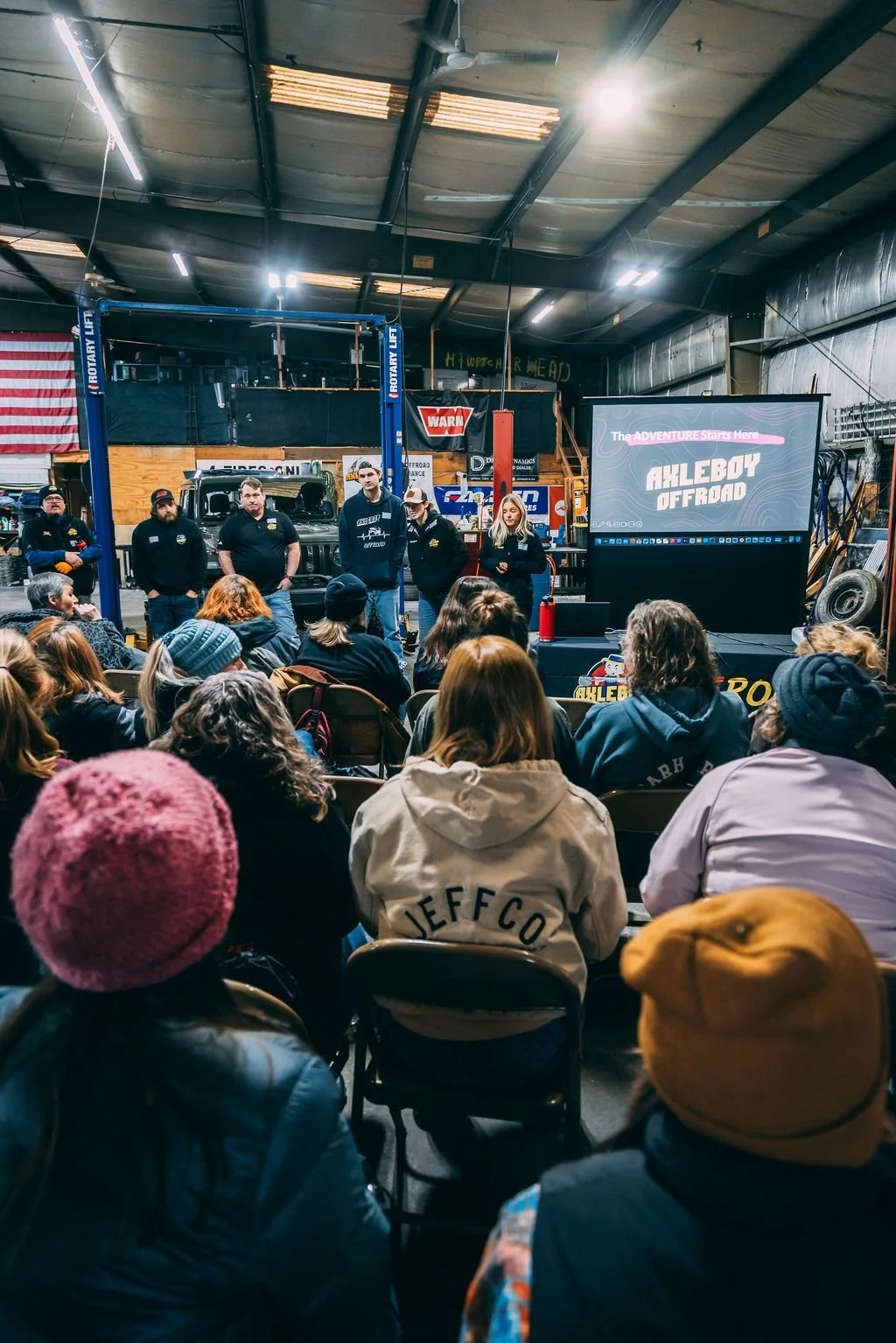 A group of people attending a presentation inside a workshop or garage. Five speakers face the audience, standing near off-road vehicles and a large screen that displays the text 'The ADVENTURE Starts Here' and 'AHLEBY OFFROAD'. The audience is seated and listening attentively.