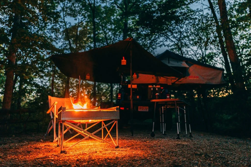 Campfire lit inside a portable fire pit, surrounded by trees and set up on gravel, with a large black umbrella and camping chairs in the background at dusk.
