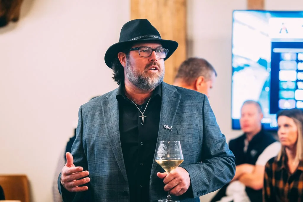 A man with glasses, gray beard, wearing a black hat and a checkered blazer, holds a glass of white wine at a social gathering, with people and a television screen in the background.