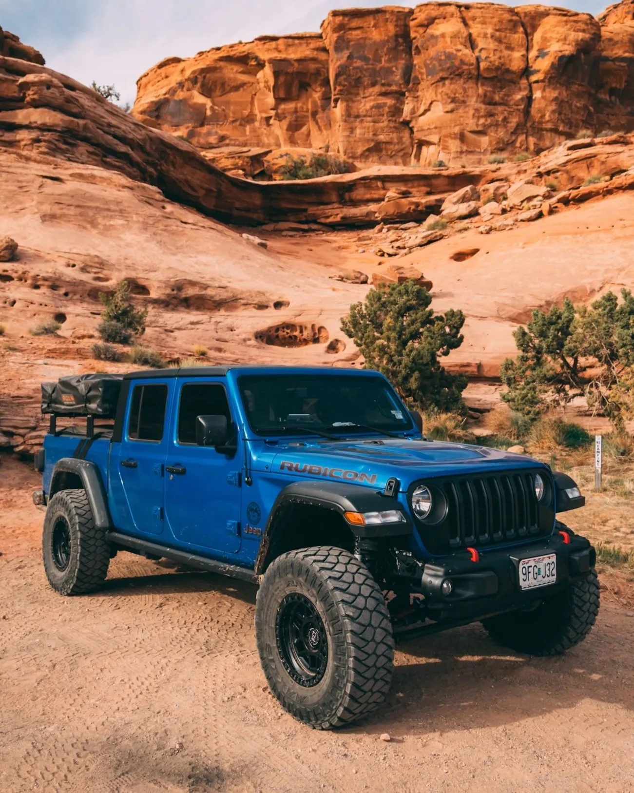 A blue Jeep Rubicon parked on a dirt trail in front of red rock formations and sparse green trees in a desert landscape.