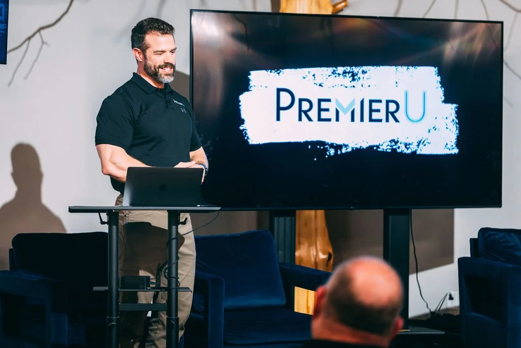 A man with a beard and black shirt giving a presentation in front of a large screen displaying the logo 'Premier U'. He is standing next to a small table with a laptop, in a room with dark blue chairs and some wall decorations.