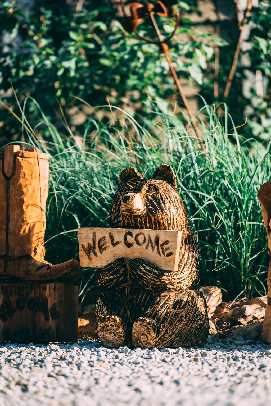 A wooden bear sculpture holding a "WELCOME" sign in front of green grass and foliage.