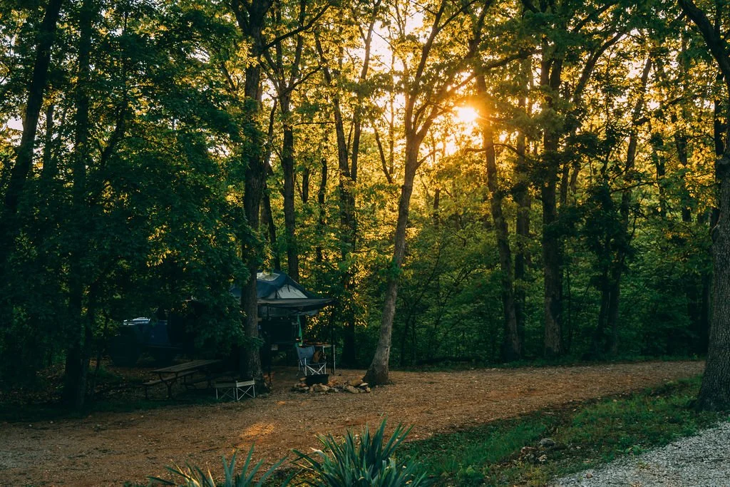 A campsite in a wooded area with a tent, camping chairs, and a picnic table, during sunset with sunlight filtering through the trees.