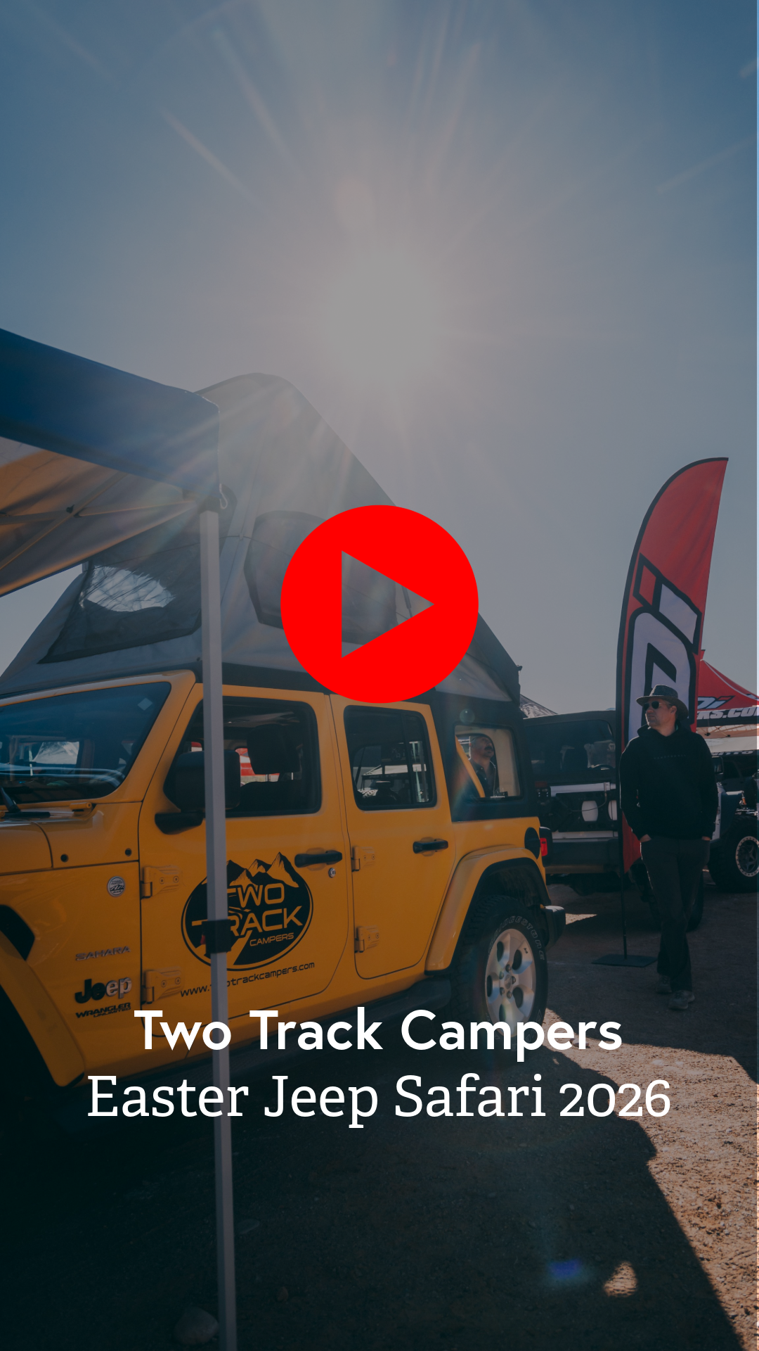 A yellow Jeep with a black and yellow "Two Track Campers" logo parked at an outdoor event with people and tents, bright sunlight, and a large red flag in the background.